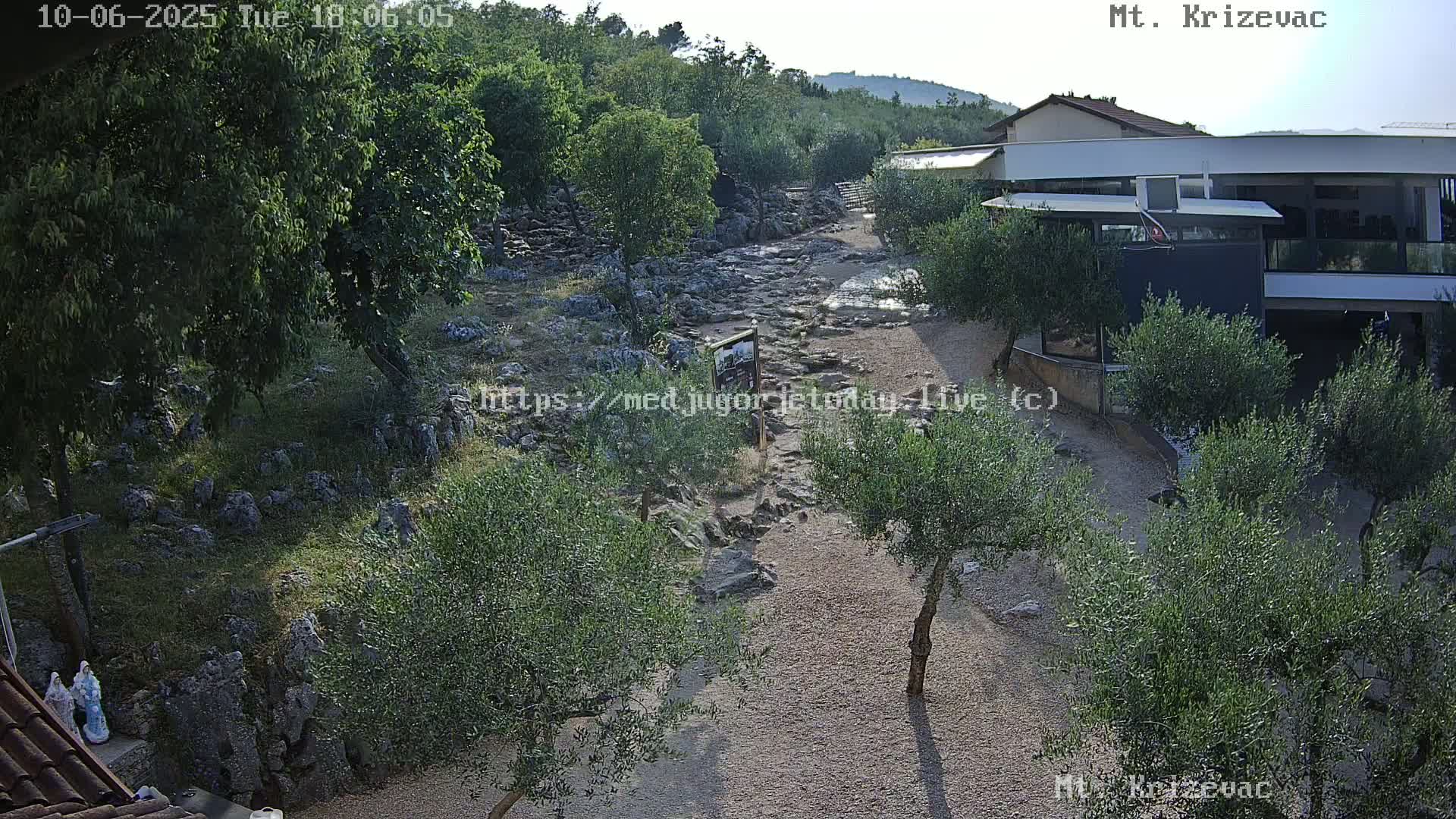 A rocky, dry riverbed winds through a landscape of olive trees and low buildings under a mostly clear sky.