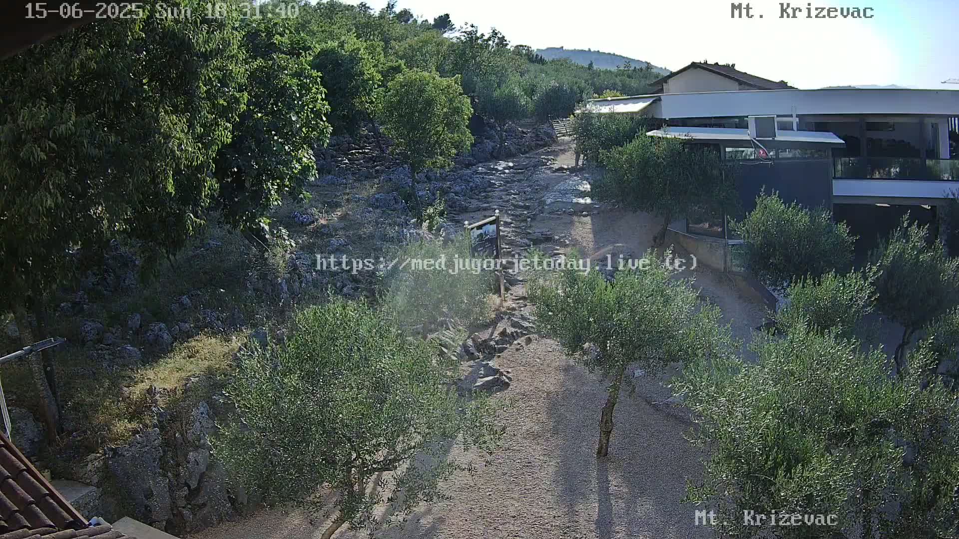 A rocky, dry creek bed winds through a landscape of olive trees and low buildings under a sunny sky.