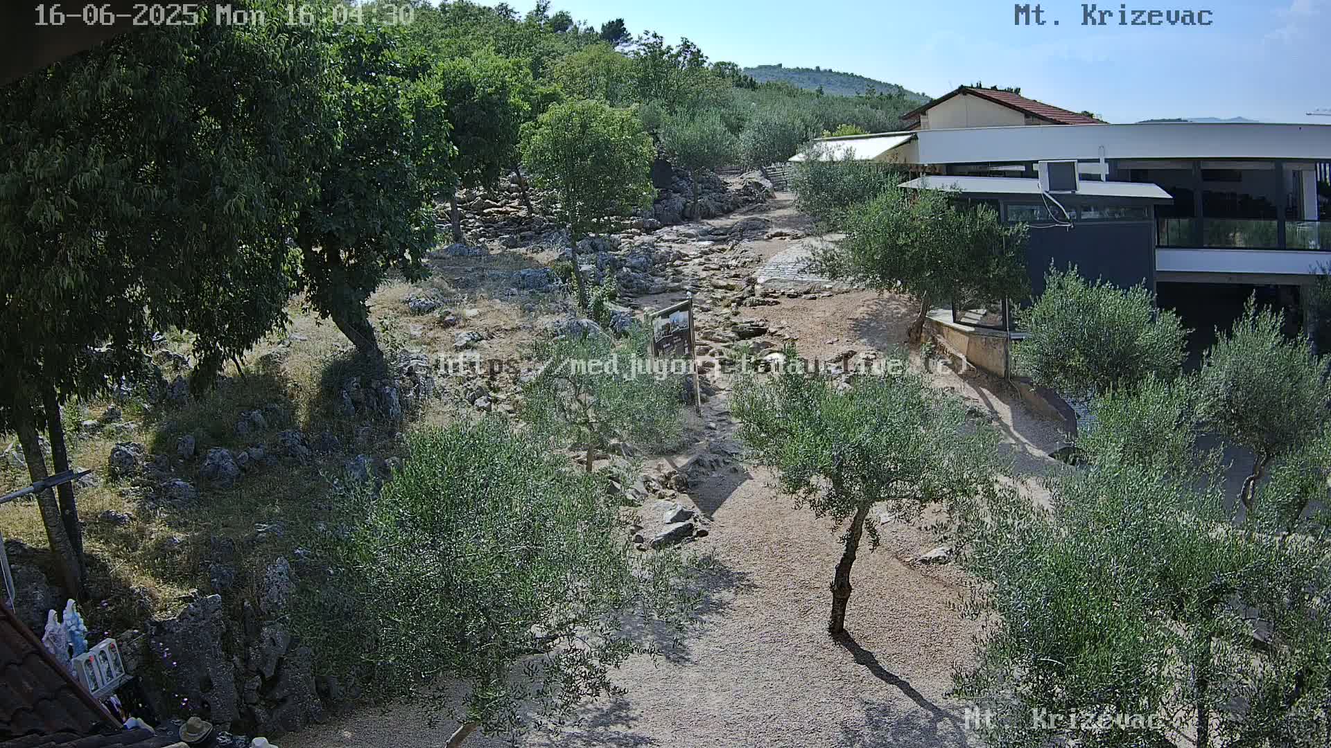 A rocky path winds through a landscape of olive trees and low shrubs under a clear, sunny sky, toward a modern building.