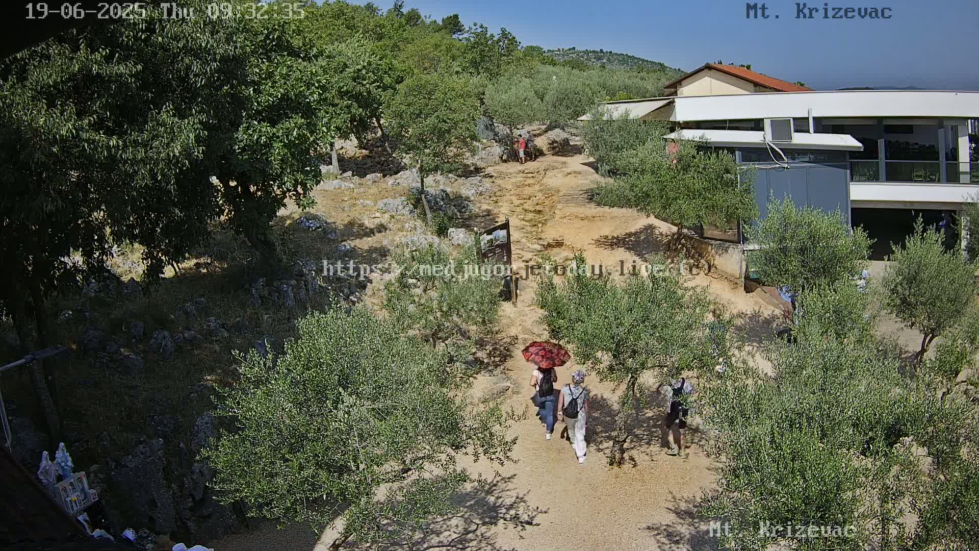 A sunny outdoor scene shows a dirt path winding through olive trees toward a building, with several people walking on the path under a red umbrella.