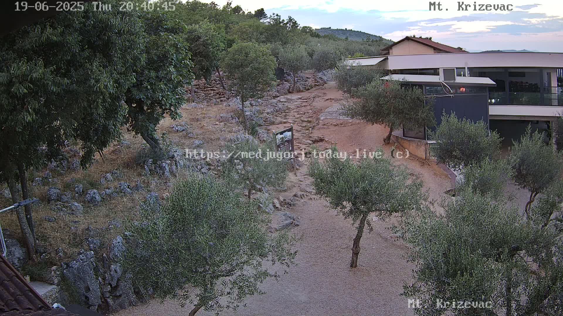 An outdoor scene under a partly cloudy sky shows a rocky path winding through a landscape of olive trees and low-lying vegetation, leading to a modern building.