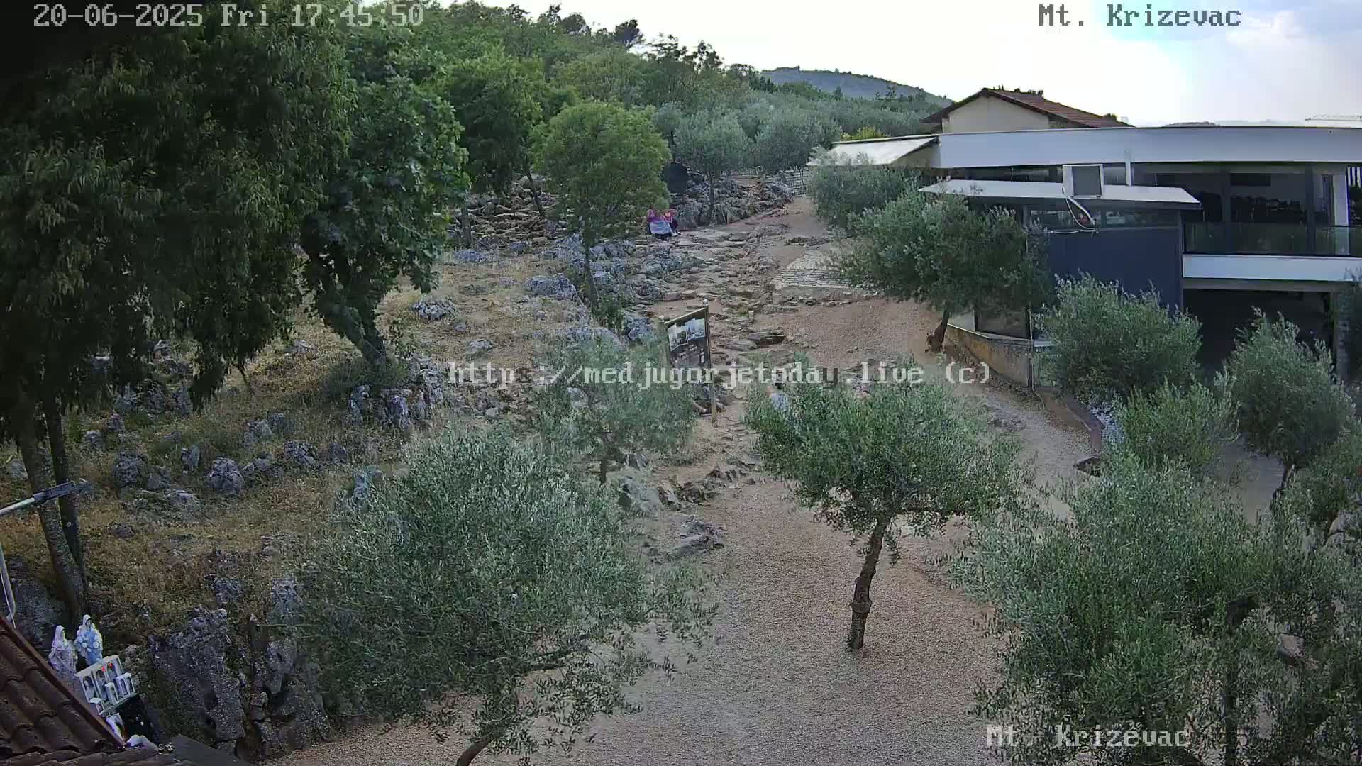 A rocky path winds through a landscape of olive trees and low buildings under a clear sky.