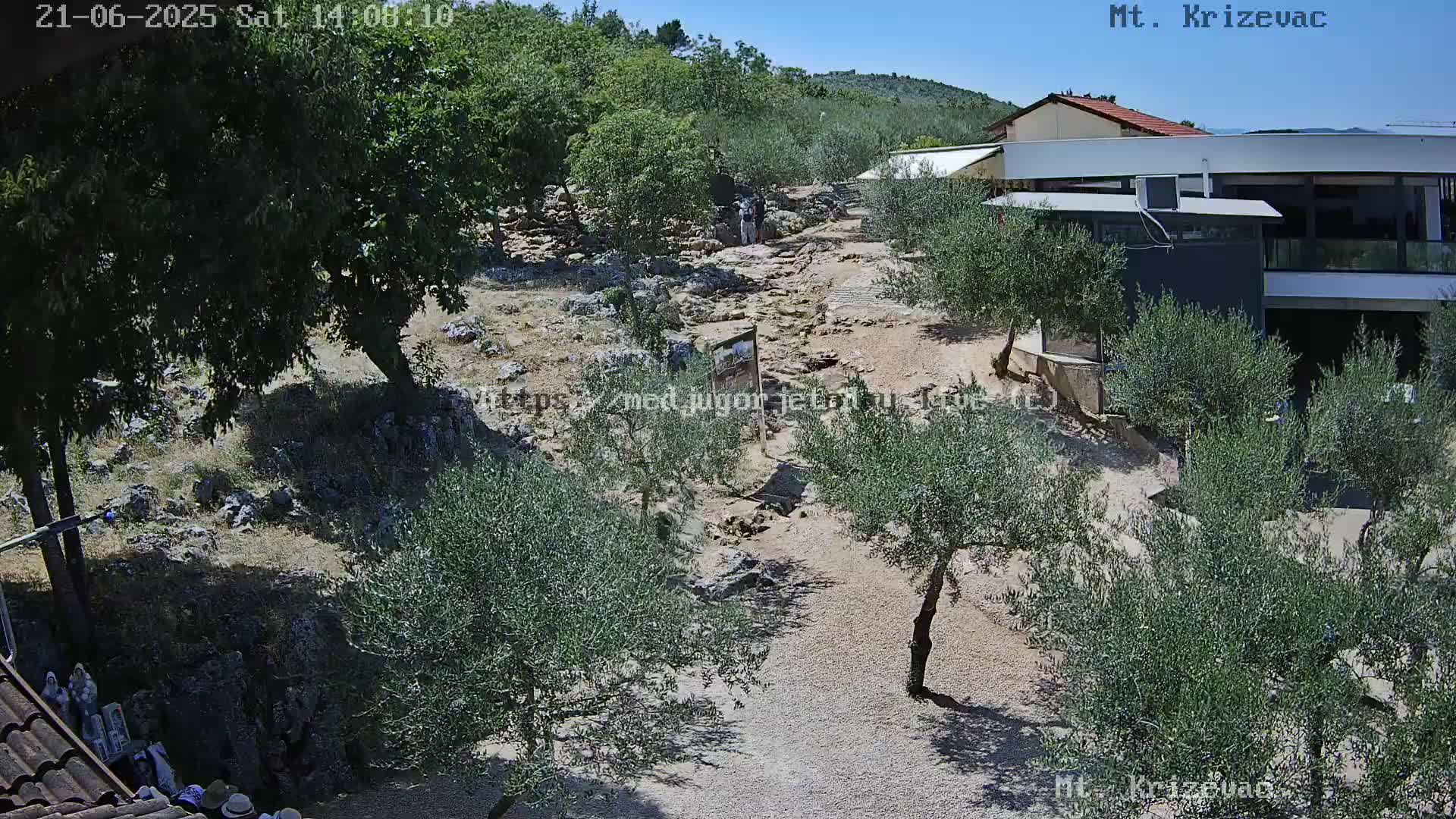 A rocky, sun-drenched landscape features olive trees, a building, and a pathway winding through the terrain under a clear blue sky.
