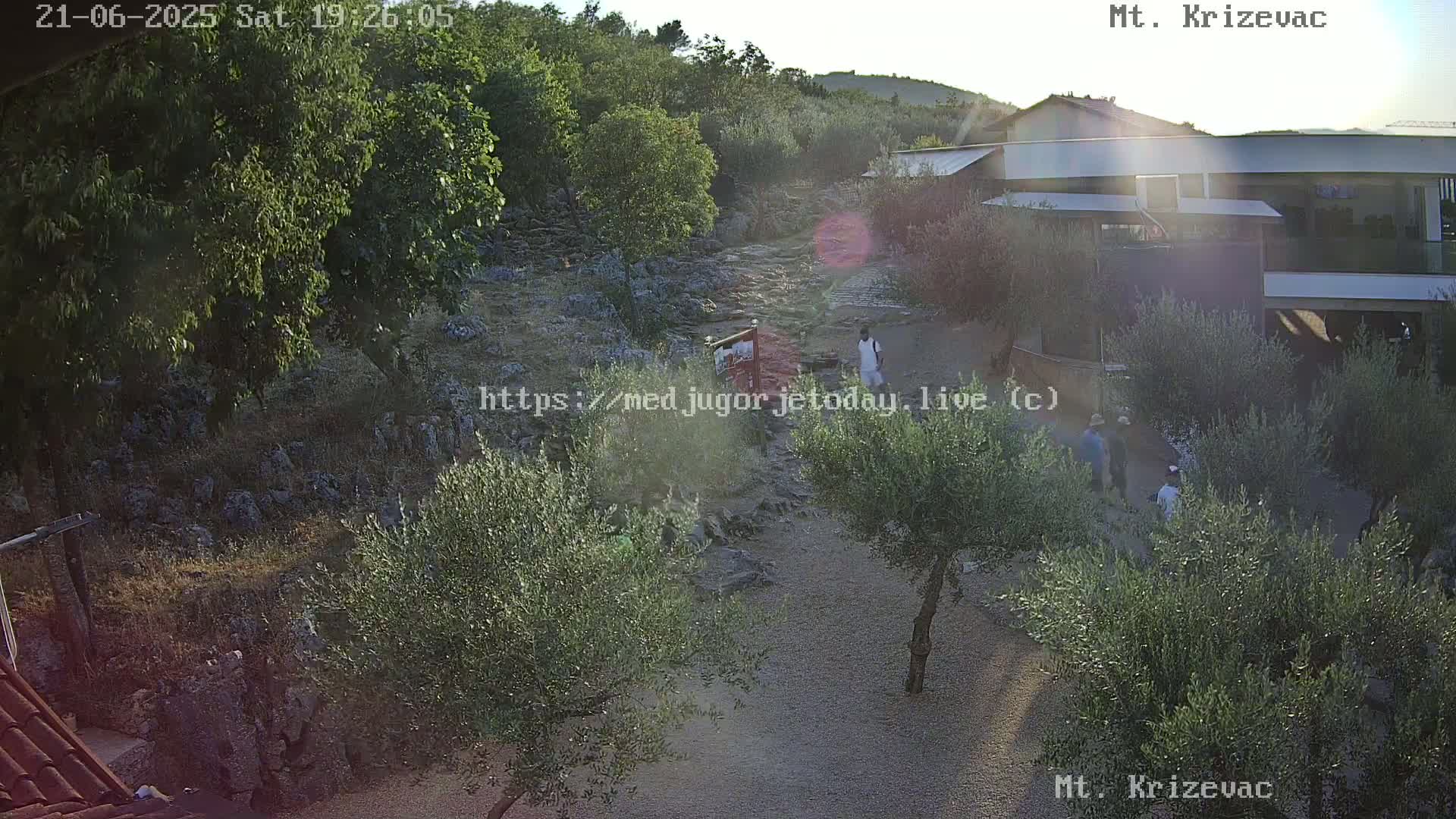 An outdoor scene at sunset shows a rocky path winding through olive trees toward a modern building, with several people walking along the path.