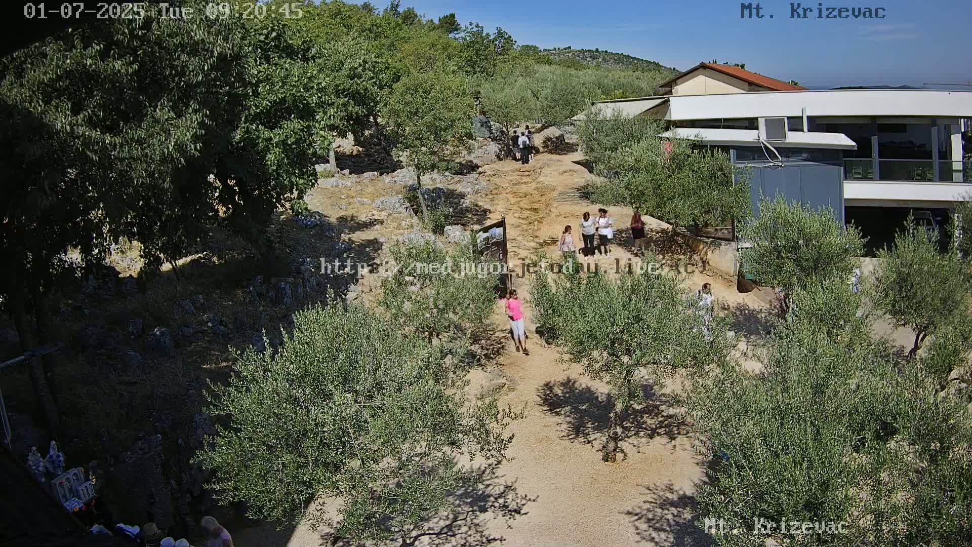 A dirt path winds through a sunny landscape of olive trees and low buildings, with several people walking along it.