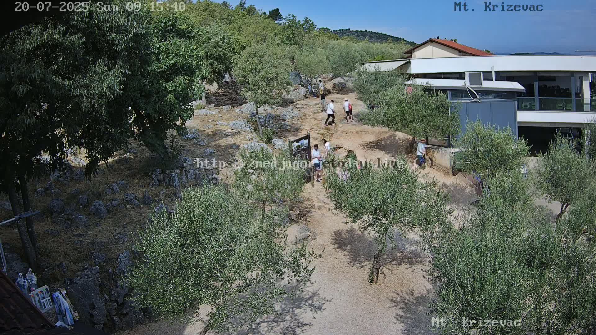 A gravel path winds through a rocky area dotted with olive trees, where several people walk near a building under a clear, sunny sky.