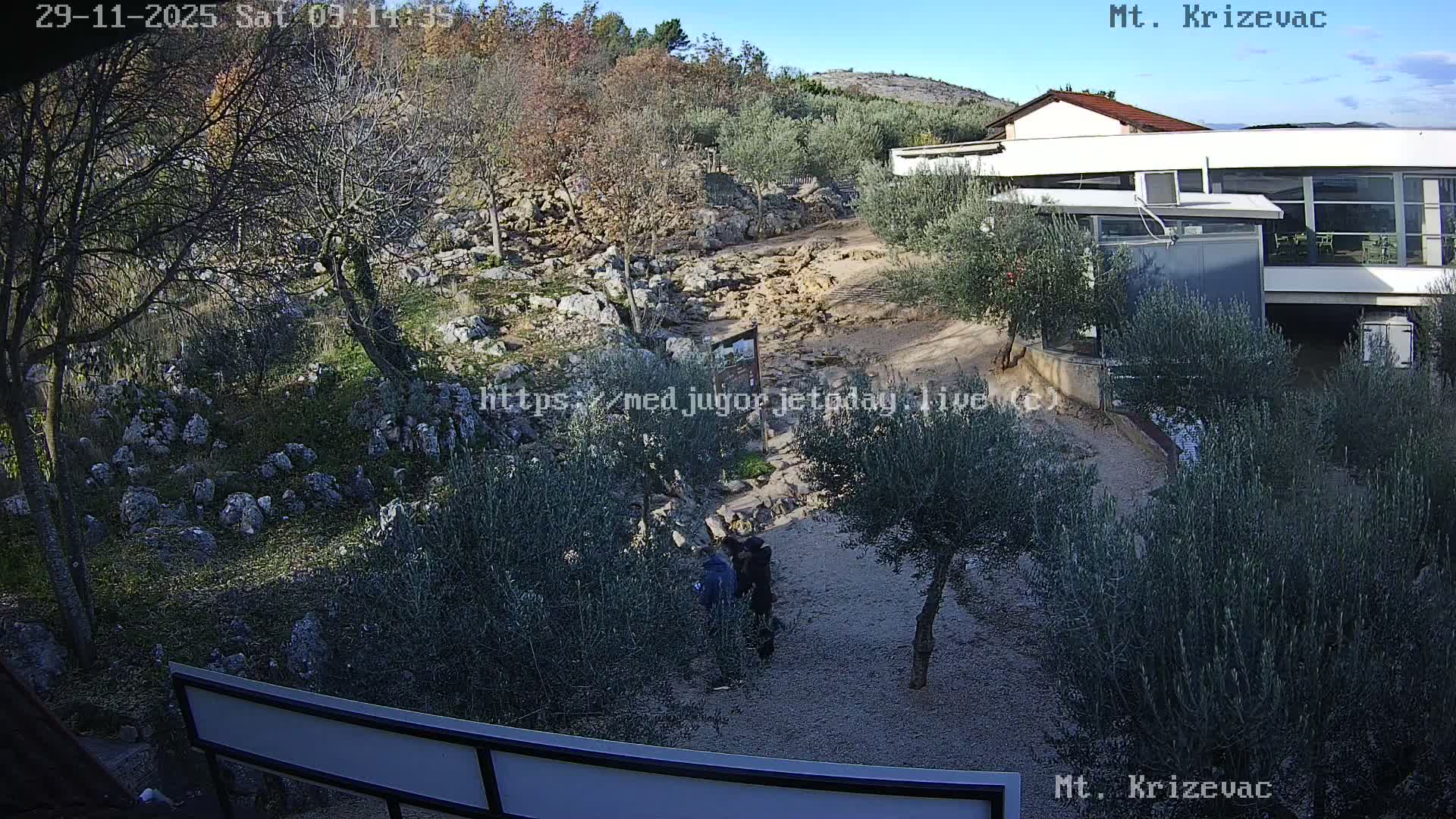 A clear and sunny day illuminates a rocky hillside with olive trees and sparse vegetation, where two people walk on a path leading past modern and traditional buildings.
