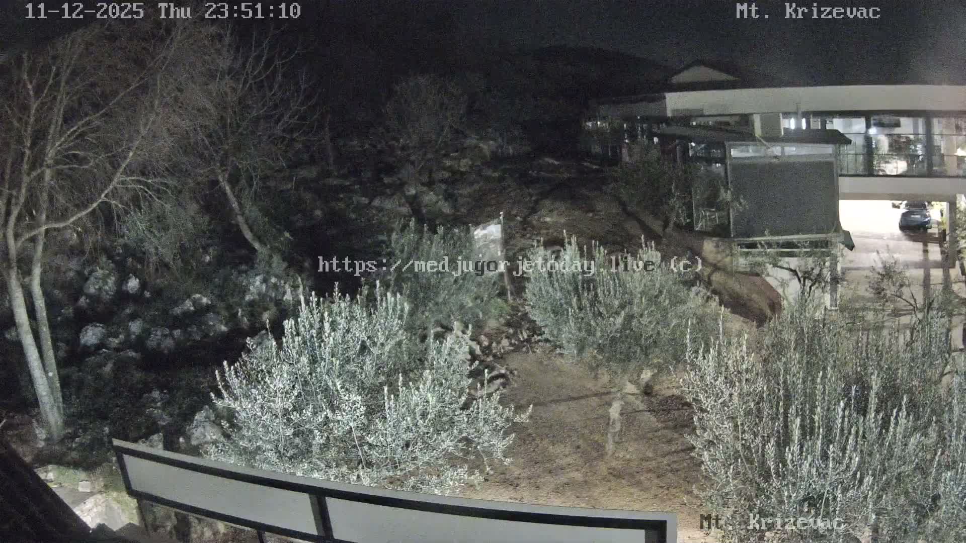 A clear night reveals a rocky, tree-covered hillside with dense olive-like bushes in the foreground, leading to a modern building with illuminated windows and a parked car on the right.