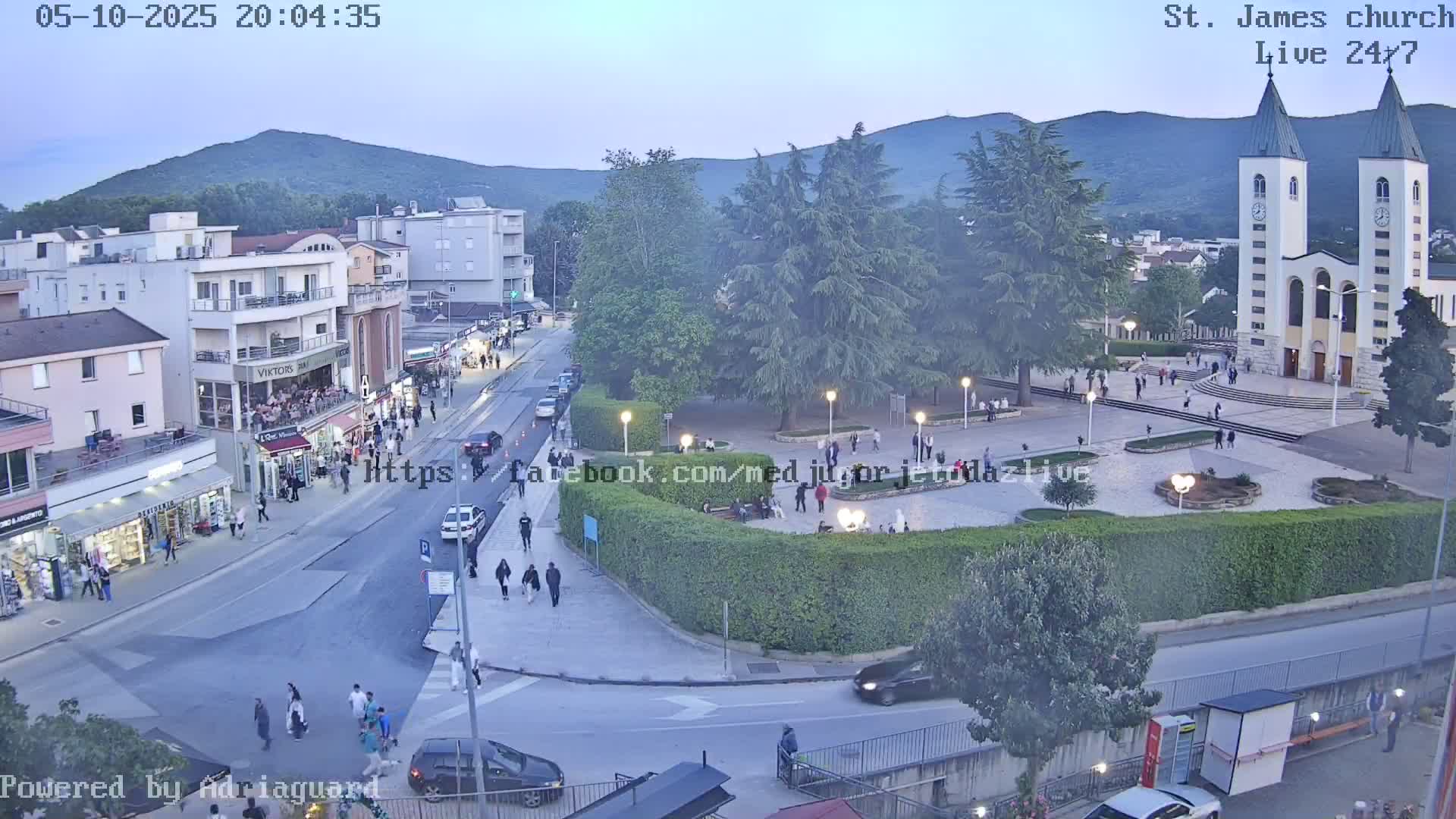 A high-angle, twilight view of a town square with a church, several buildings, pedestrians, and cars, under a mostly clear sky.