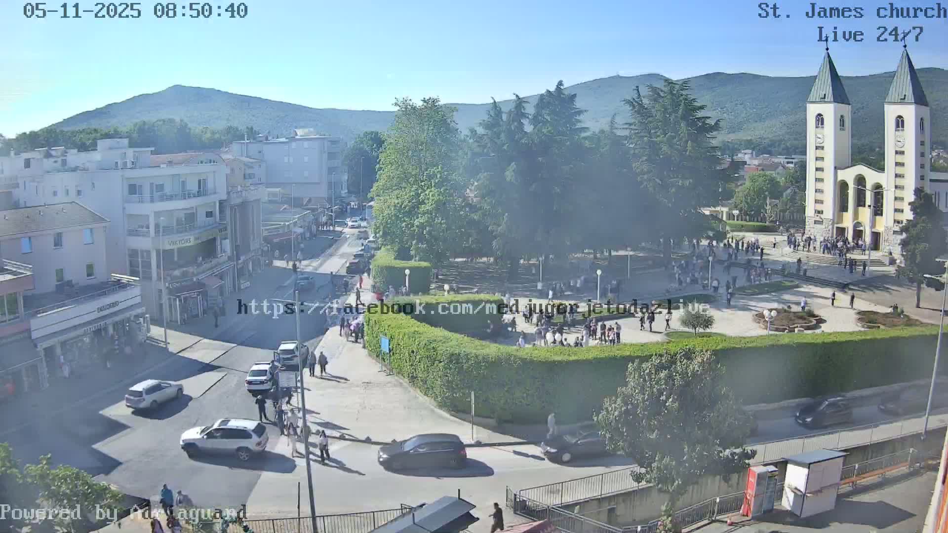 A sunny town square with a large church, numerous people, and several cars is viewed from above.