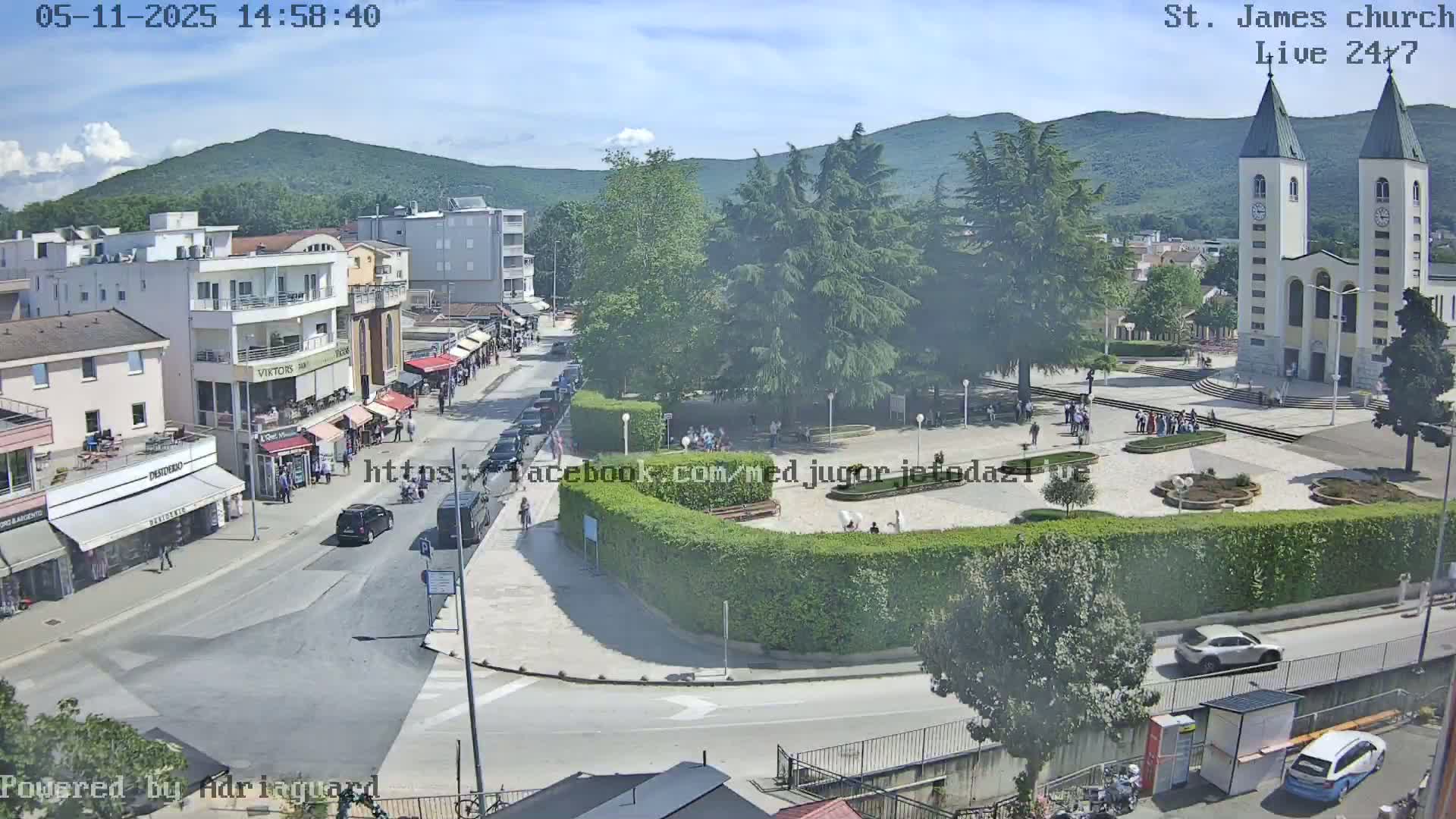 A high-angle view shows a town square with a church, buildings, cars, and people on a sunny day.