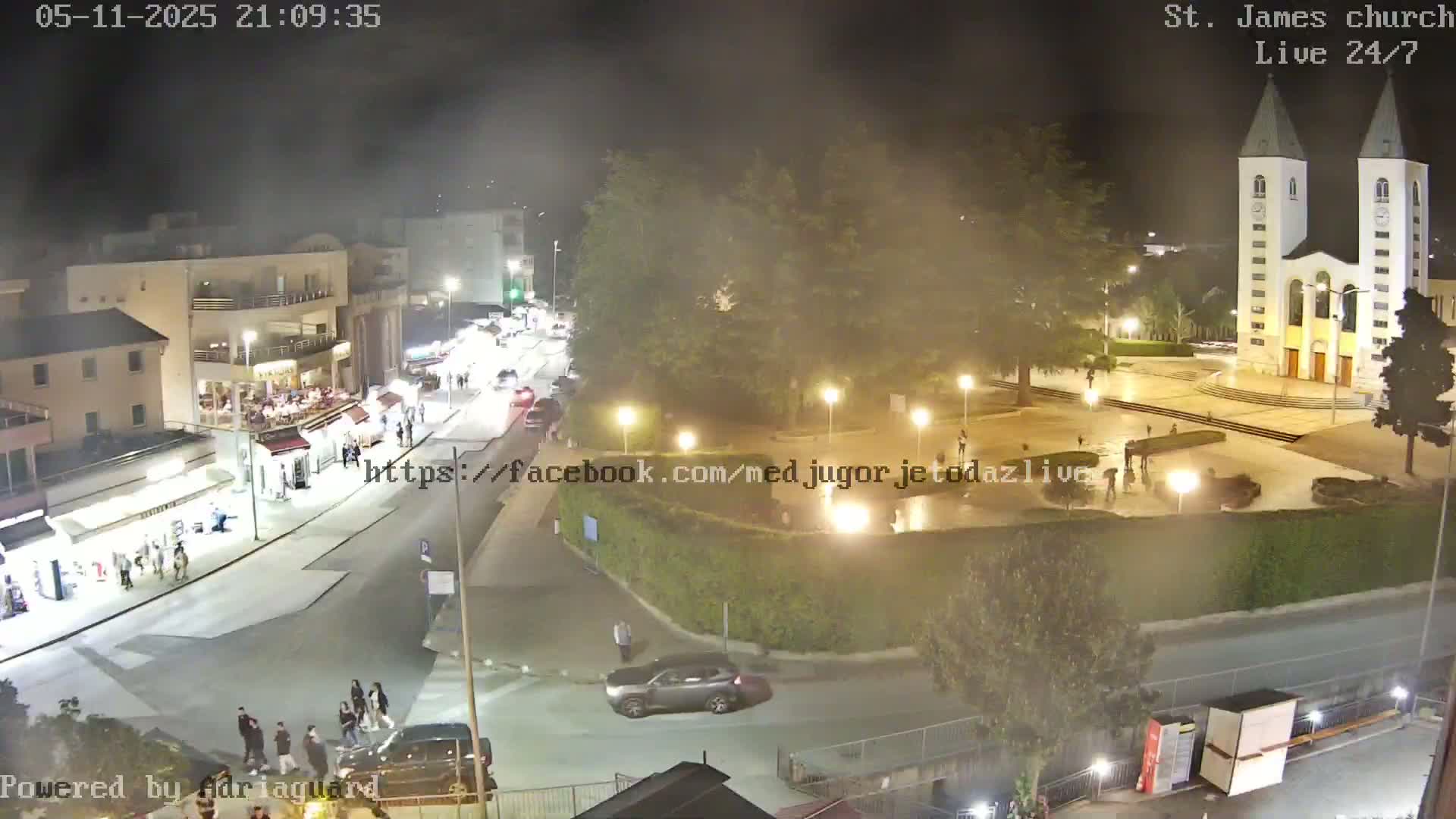 A nighttime aerial view of a town square with a church, pedestrians, and cars, under hazy or foggy conditions.