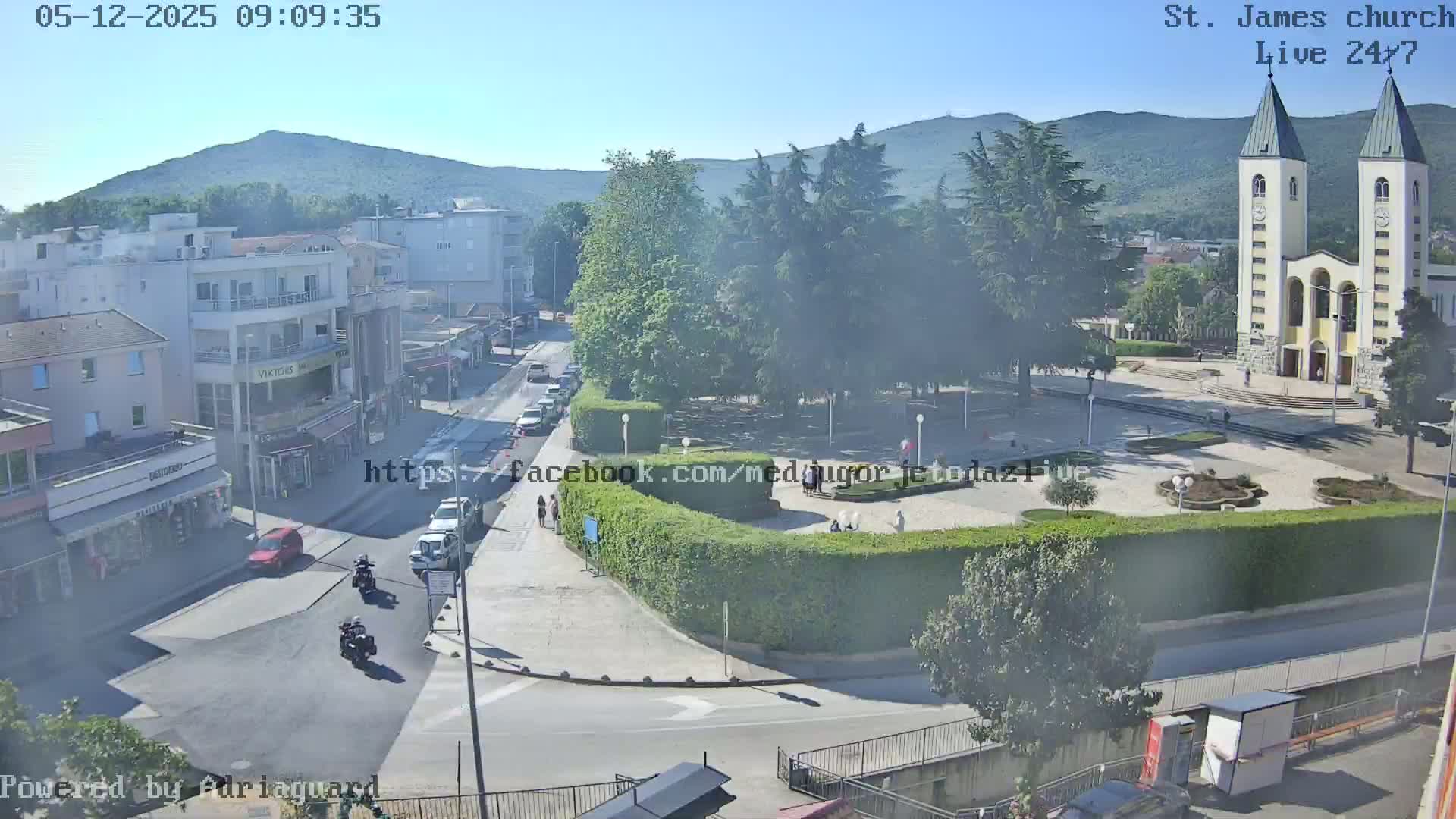 A high-angle, long shot on a sunny day shows a town square with a large church in the background, vehicles on roads, and people walking around the square.