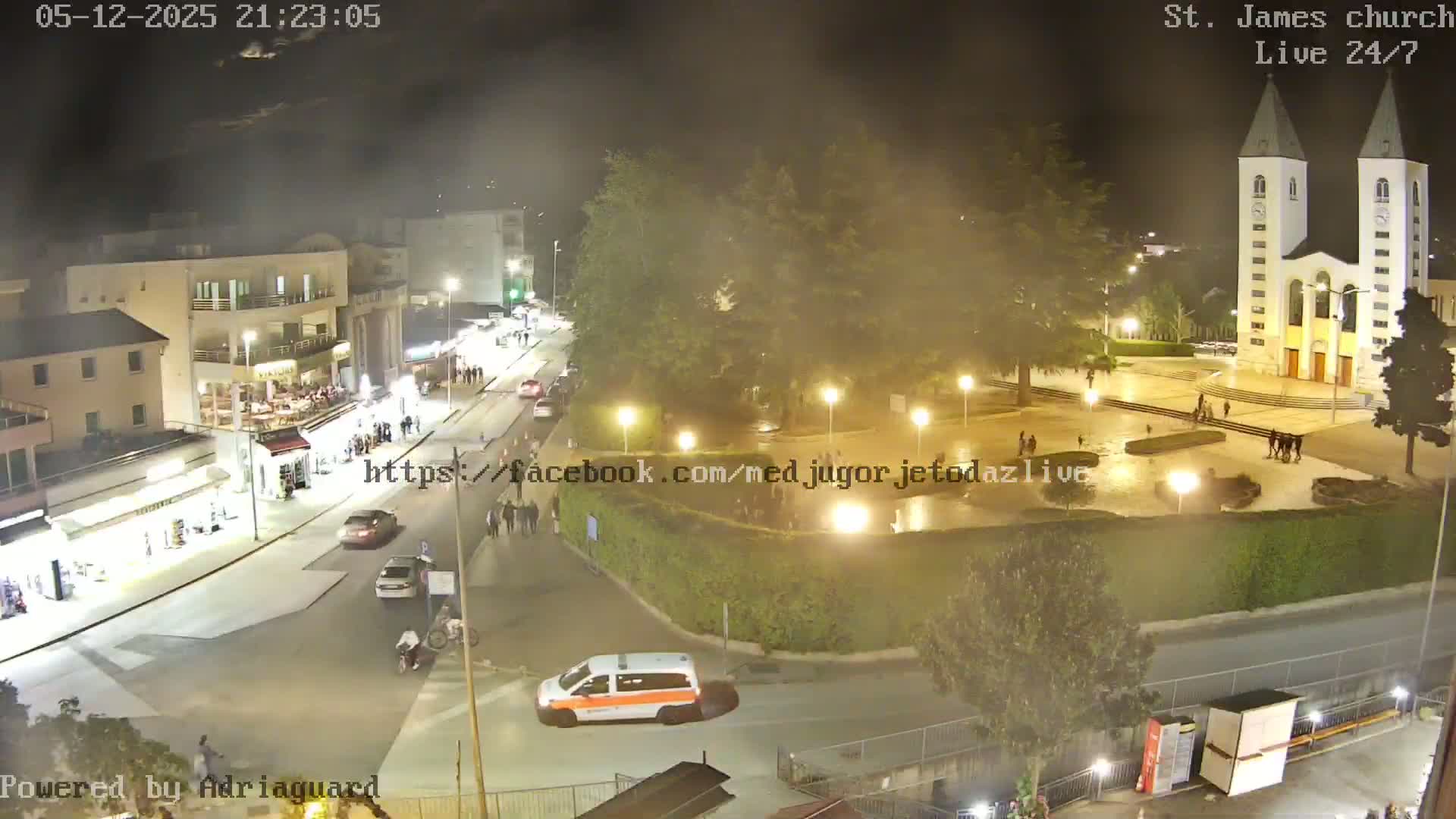 A nighttime aerial view of a town square with a church, some vehicles, and several people walking around under hazy conditions.