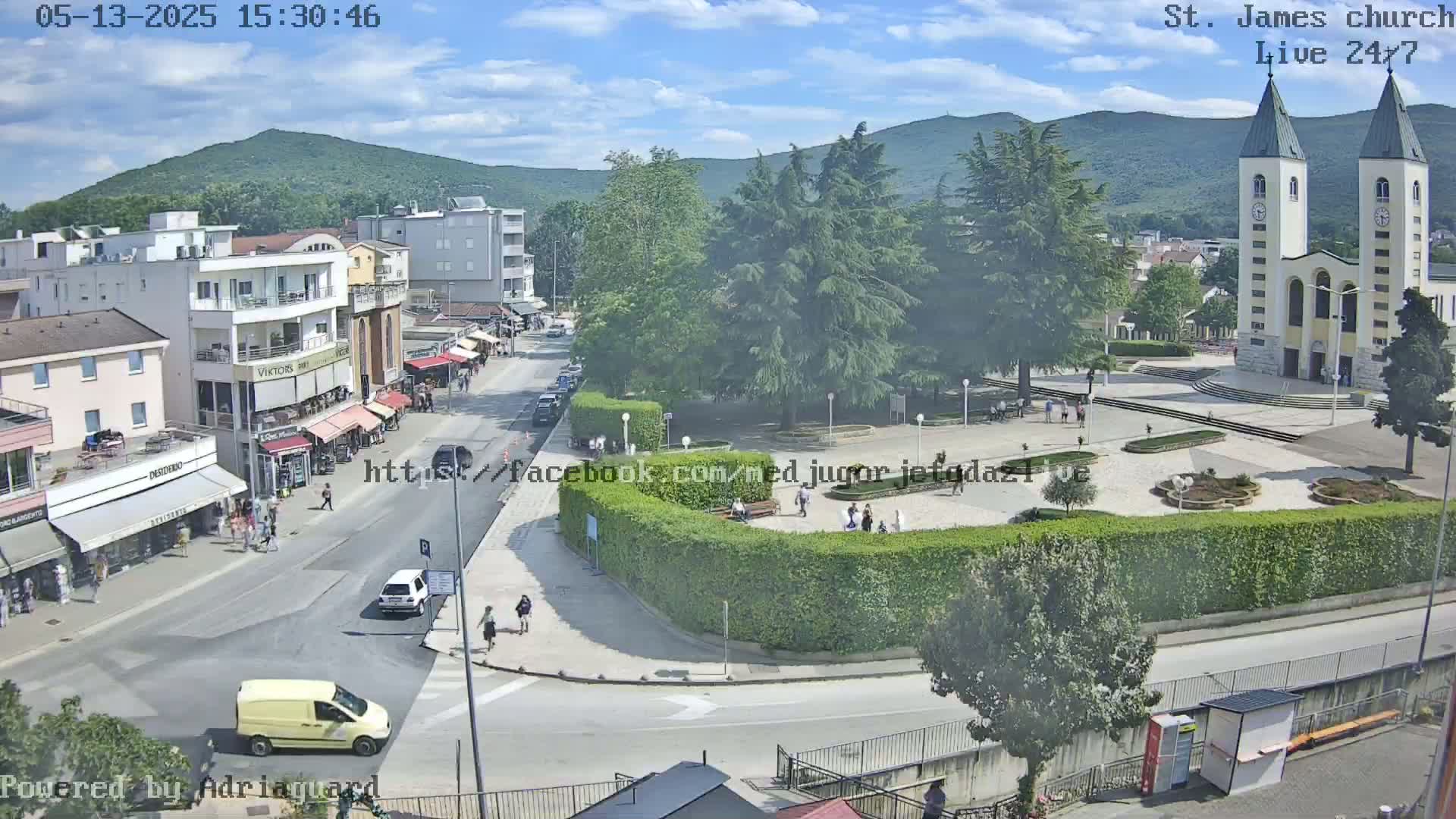 A town square with a church, shops, and people walking around on a sunny day, with mountains visible in the background.