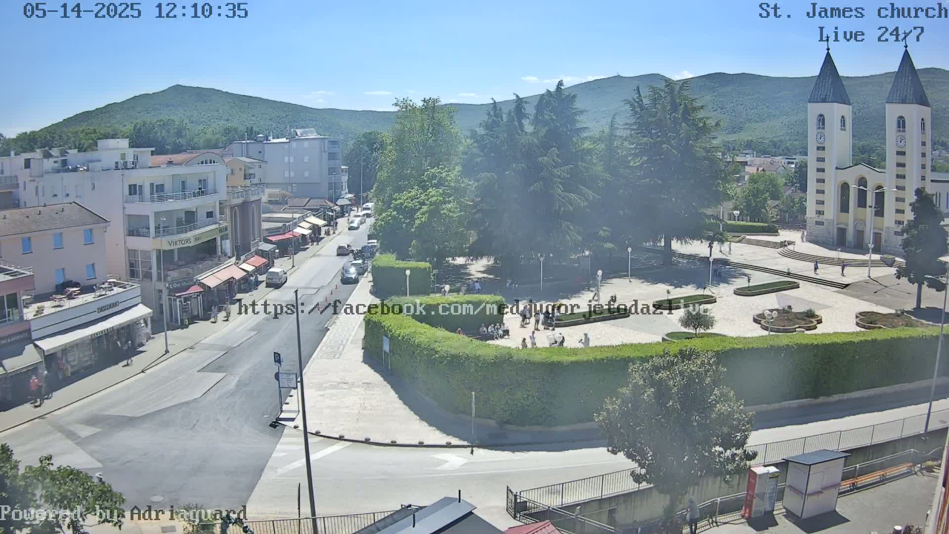 A sunny day view of a town square with a church, buildings, and people, surrounded by lush greenery and mountains in the background.