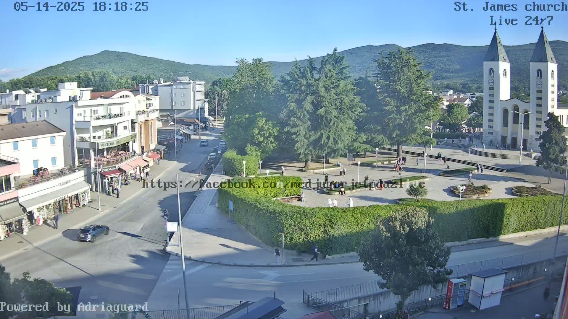 A sunny day shows a town square with a church, several buildings, cars, and people in a plaza, all surrounded by green trees and hedges.