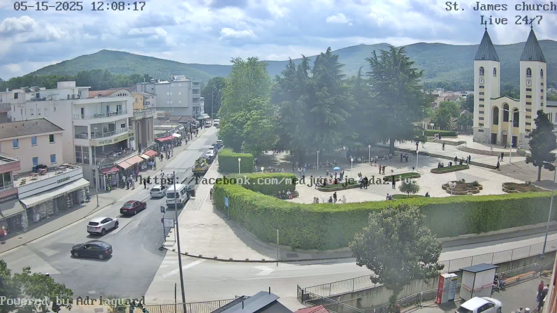 A partly cloudy day reveals a town square with a church, surrounding buildings, and several cars parked and driving on streets; many people are visible in the square and streets.