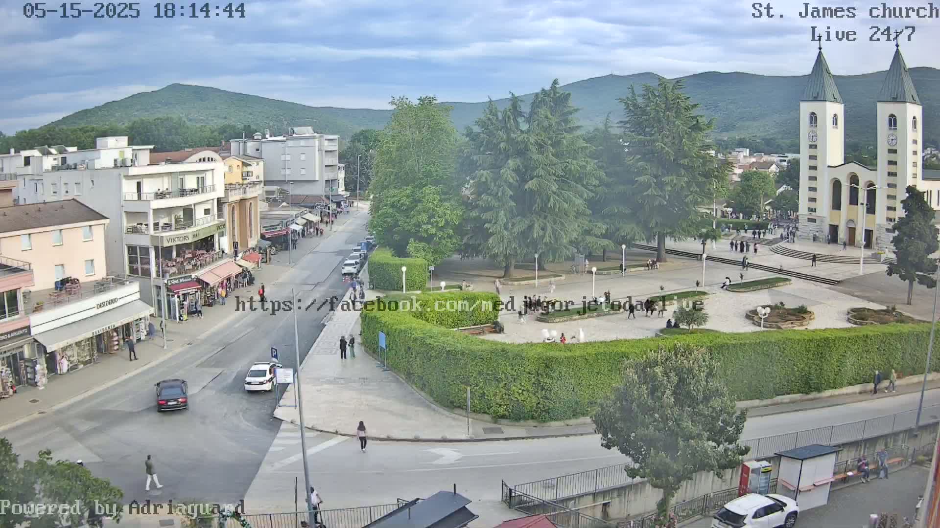 An overcast day view of a town square with a church, buildings, cars, and pedestrians, surrounded by green trees and hills in the background.