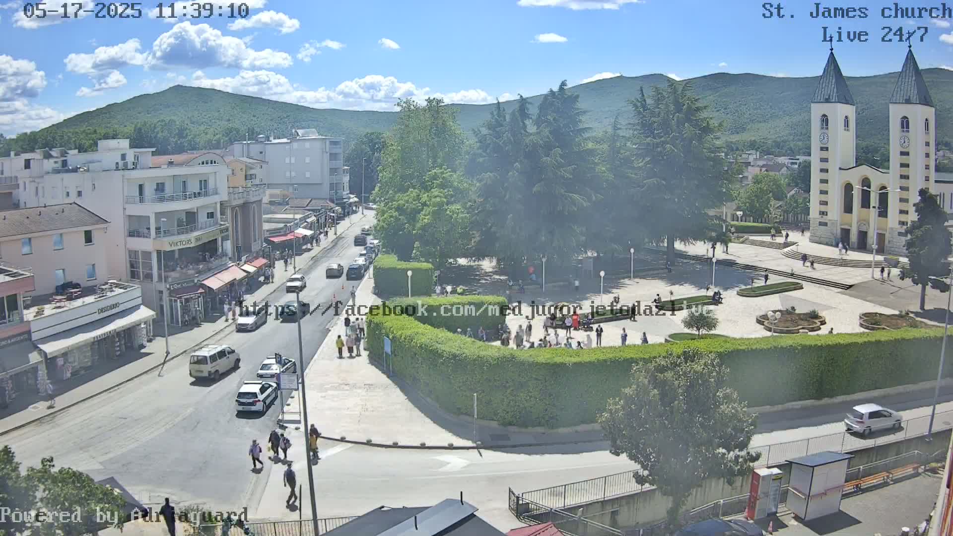A sunny day reveals a town square with a church, cars on a street, and people gathered in the plaza, with mountains in the background.