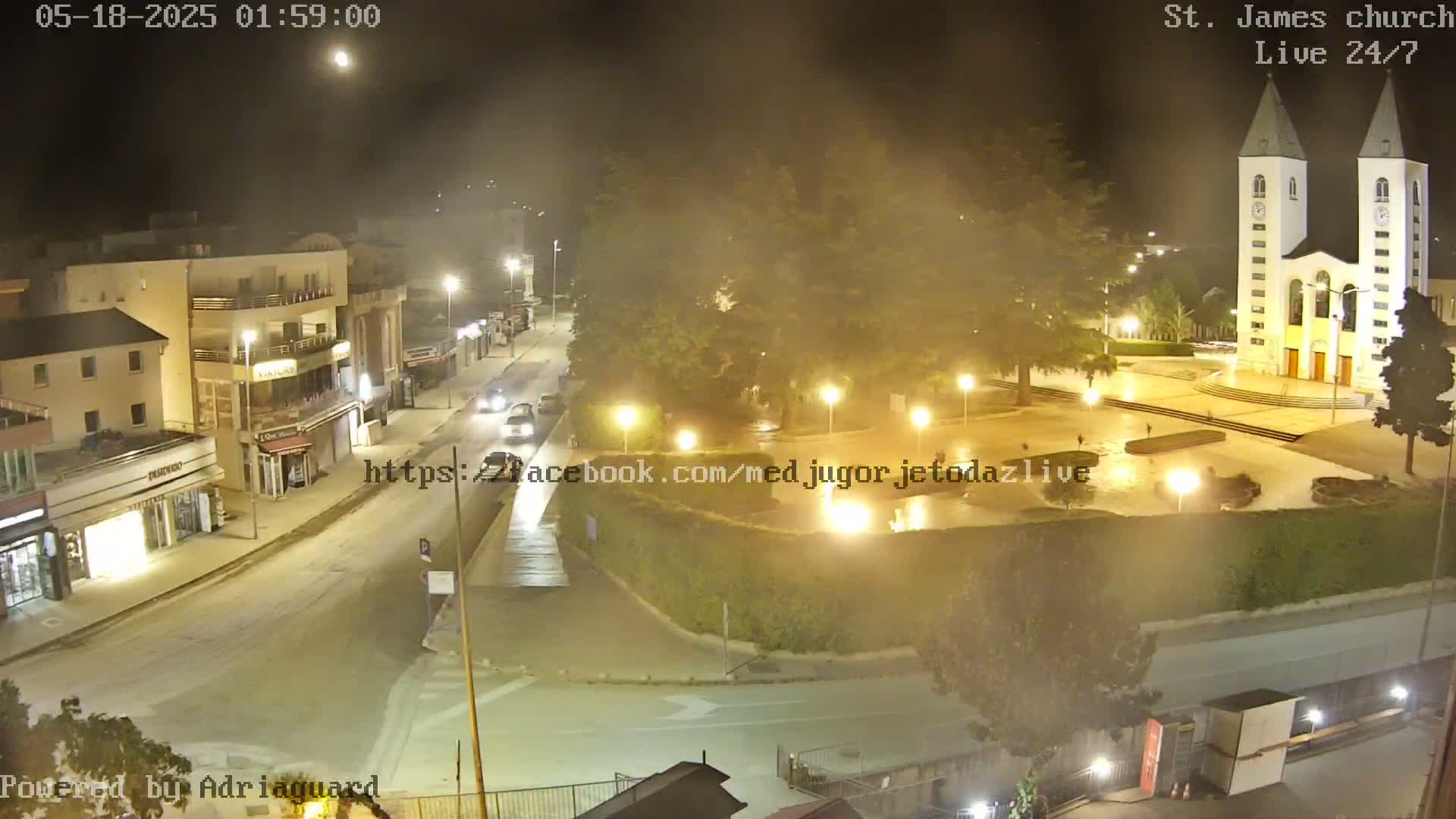 A nighttime, hazy view of a town square with a church, buildings, and cars; some streetlights illuminate the scene.