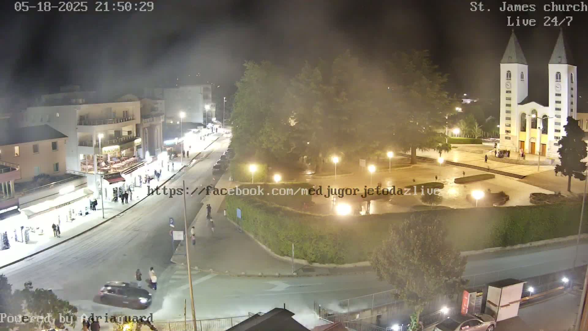 A nighttime, hazy view of a town square with a church, several buildings, and cars, with people walking around.