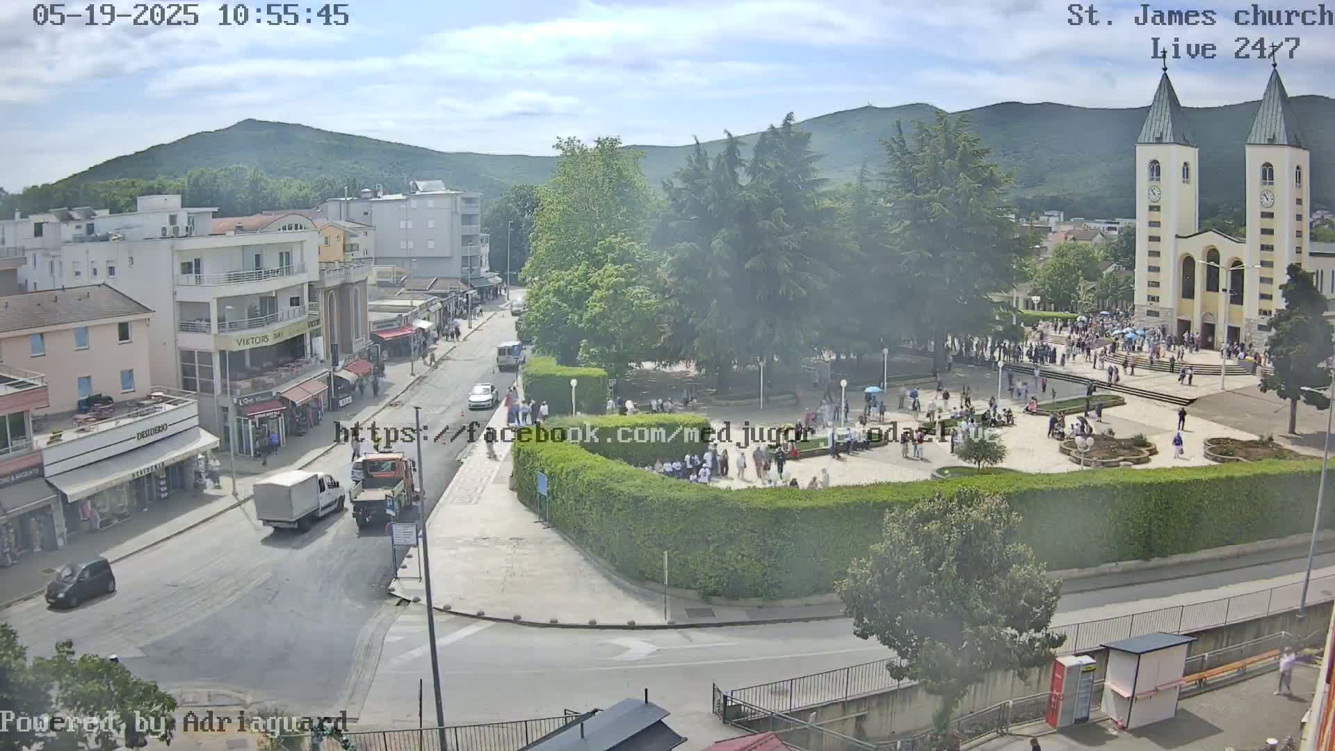 A high-angle, long shot shows a town square with a church, surrounded by buildings and people on a partly cloudy day.