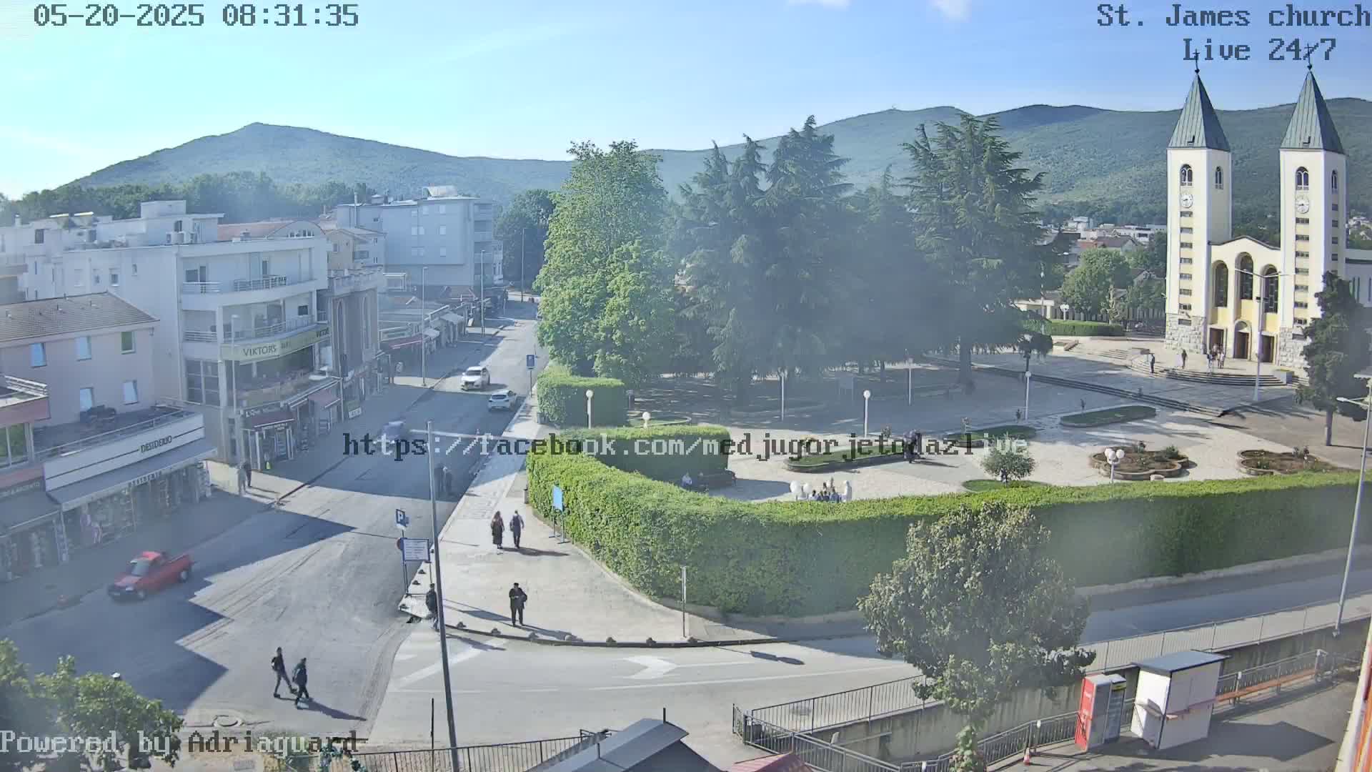 A high-angle, long shot of a town square on a sunny day, featuring a church with two tall spires and people walking around.