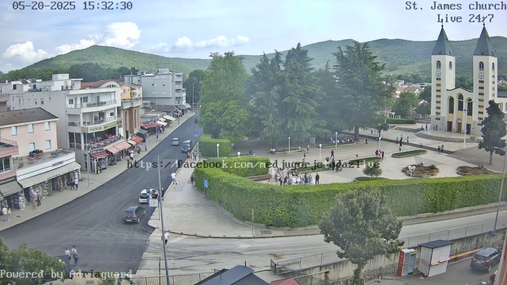 An aerial view of a town square with a church, buildings, people, and cars on a partly cloudy day.