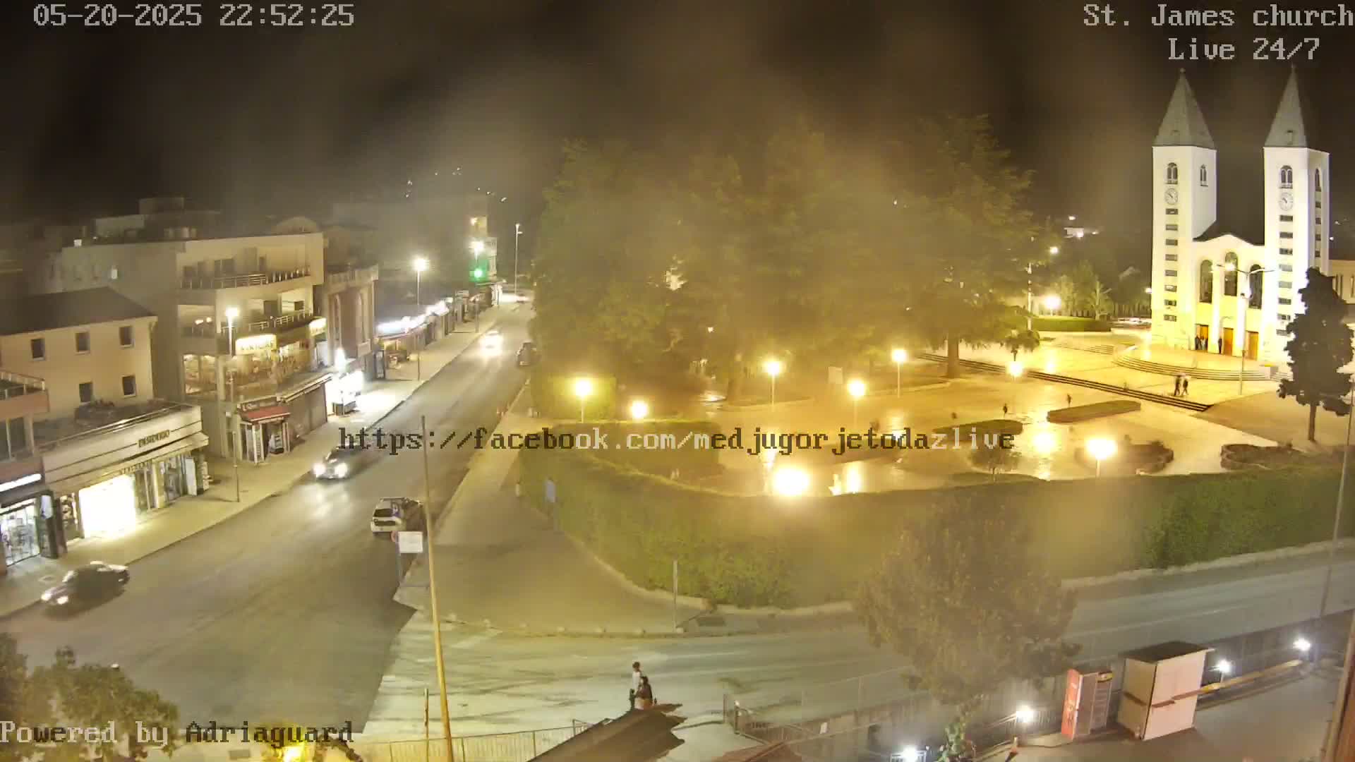 A nighttime, hazy view of a town square with a church, streetlights, and a few vehicles on the road.