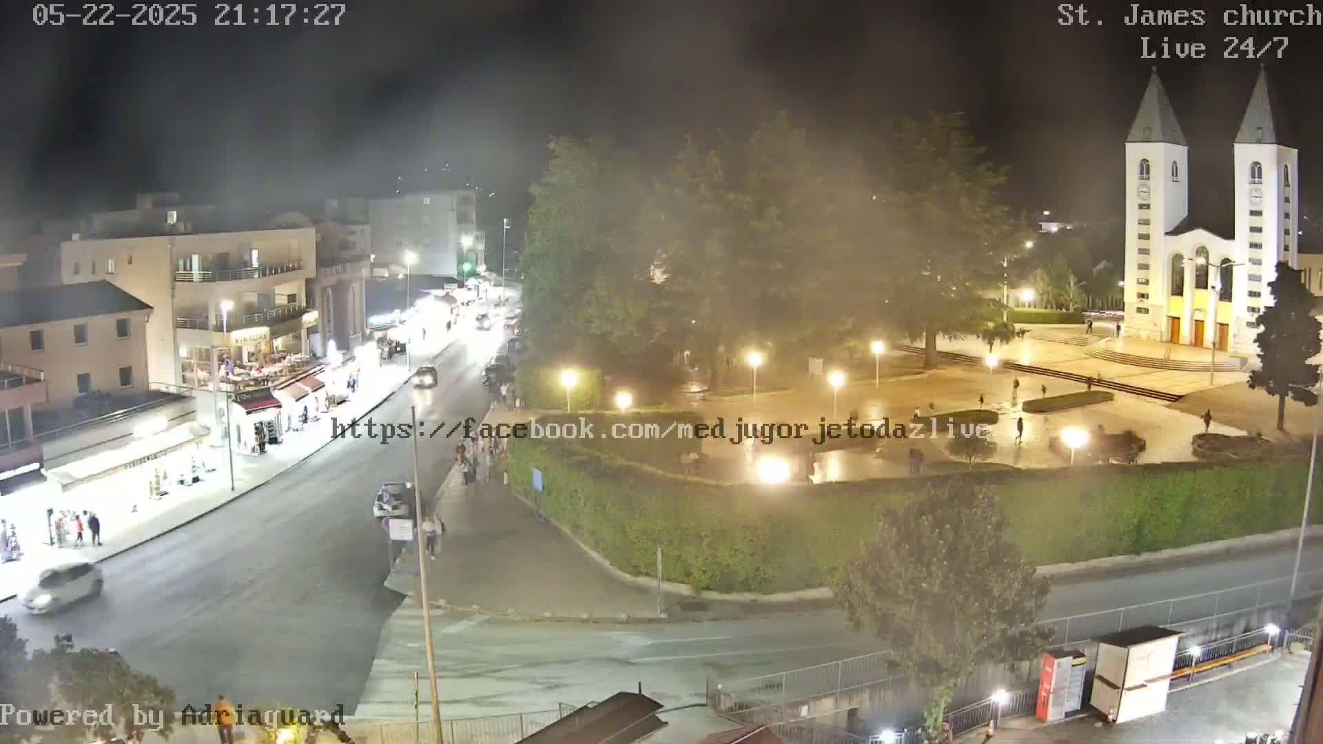 A nighttime, slightly hazy view of a town square with a church, buildings, and a street with cars and people.