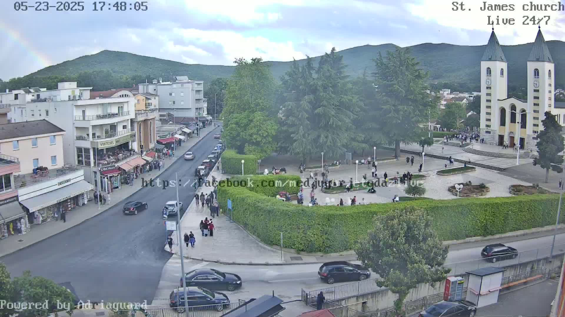 A high-angle, long shot shows a town square with a church, many people, and cars on a partly cloudy day.