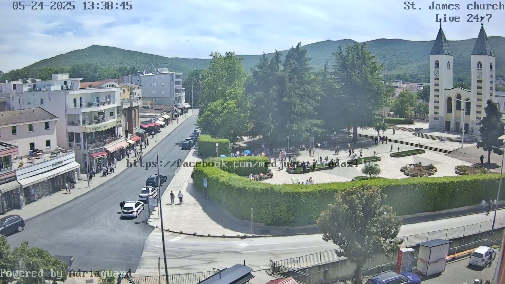 An aerial view of a town square on a sunny day, showing a church, buildings, people, and vehicles.