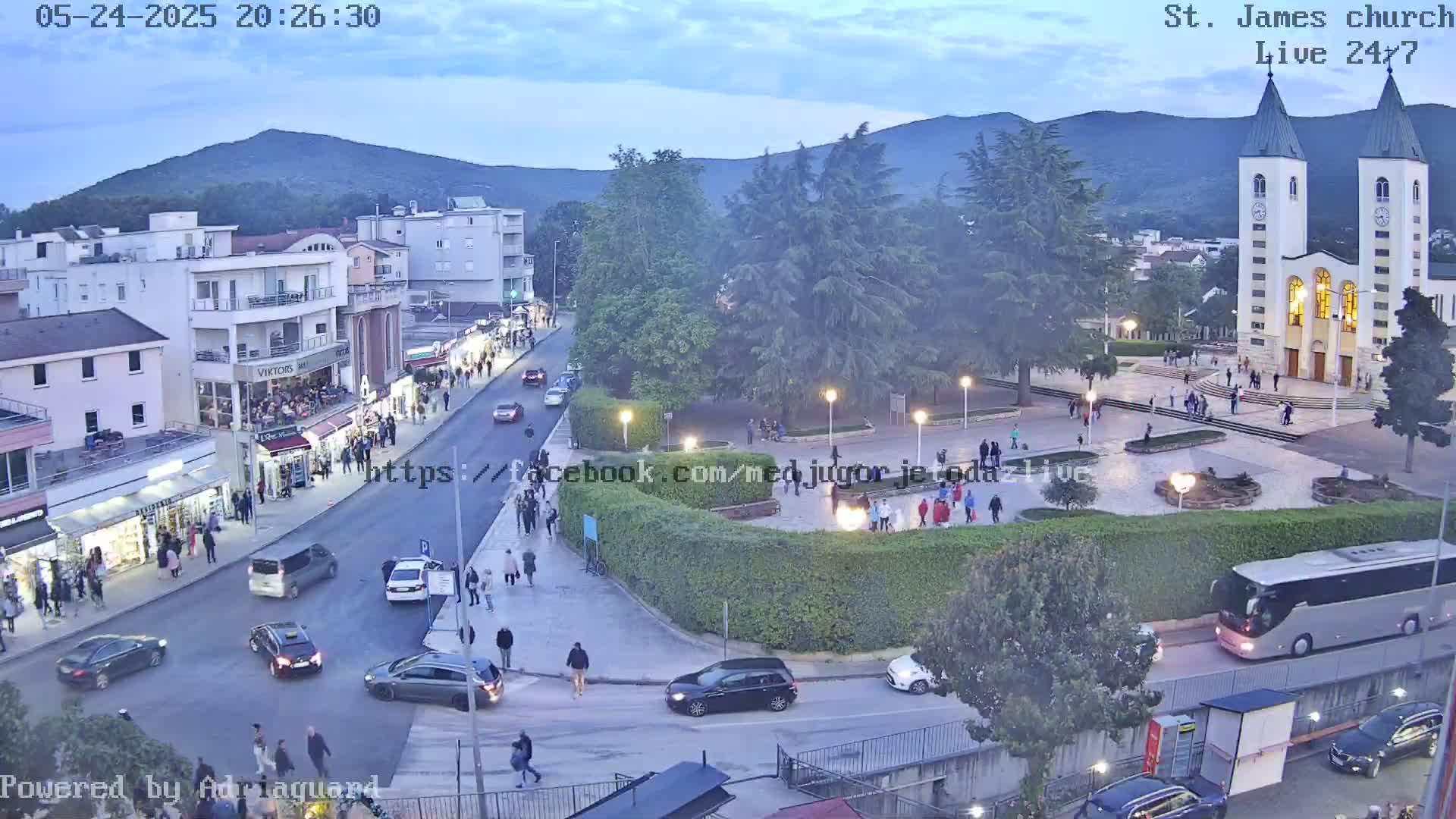 A twilight view of a town square with a church, pedestrians, and cars, under a mostly clear sky.