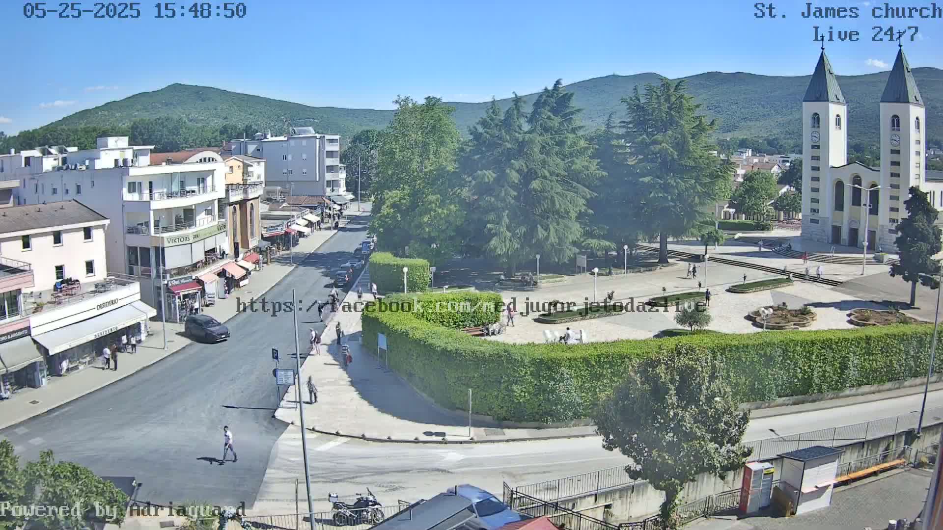 A sunny day in a town square, featuring a church, buildings, cars, people, and lush green landscaping.