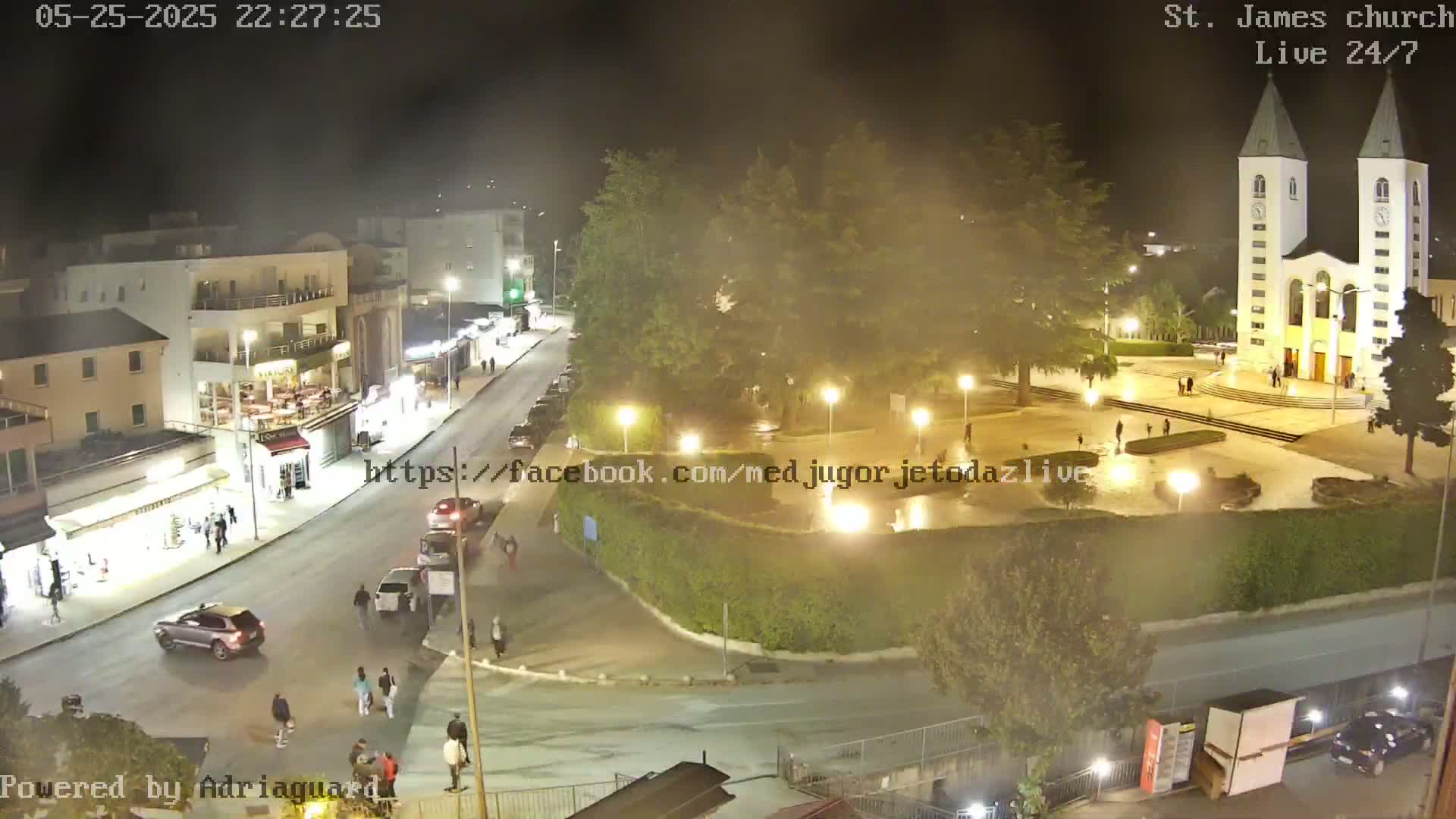 A nighttime, slightly hazy view of a town square with a church, streetlights, cars, and pedestrians.
