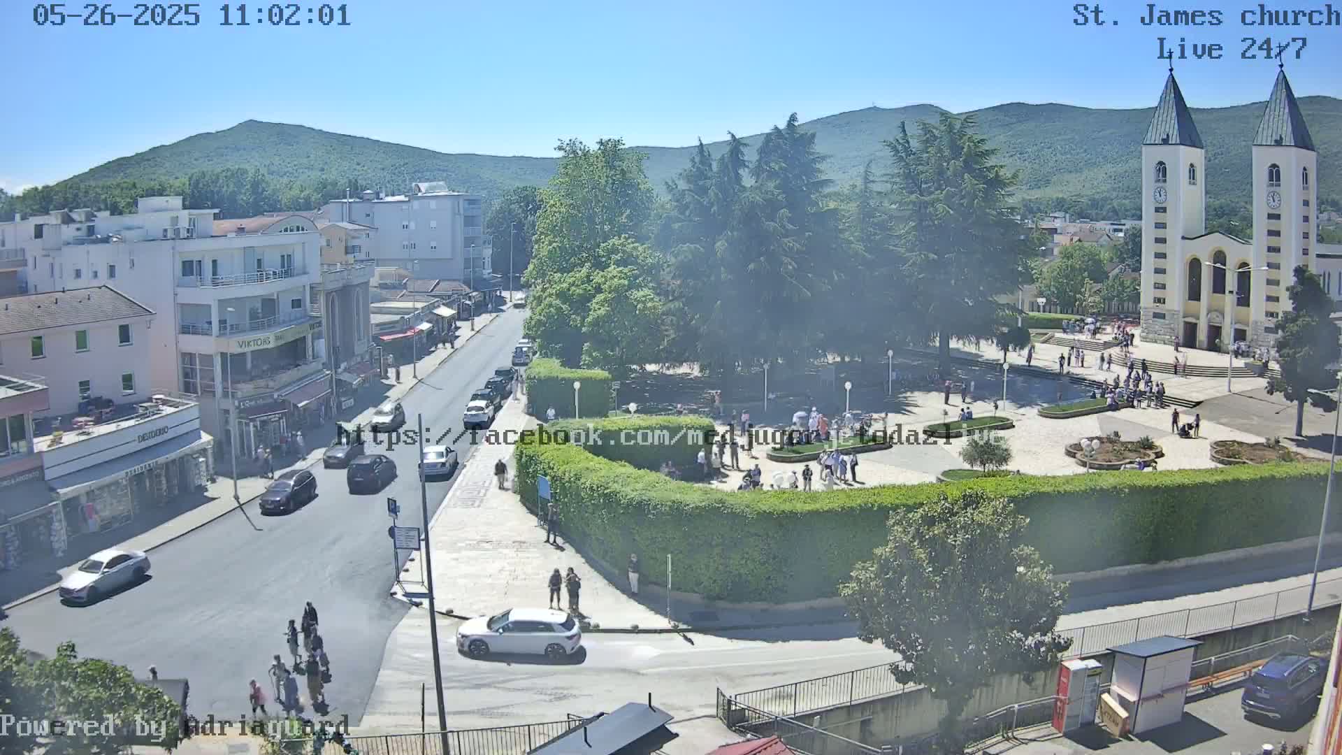 A sunny day shows a town square with a church, cars on roads, people walking around, and green hills in the background.