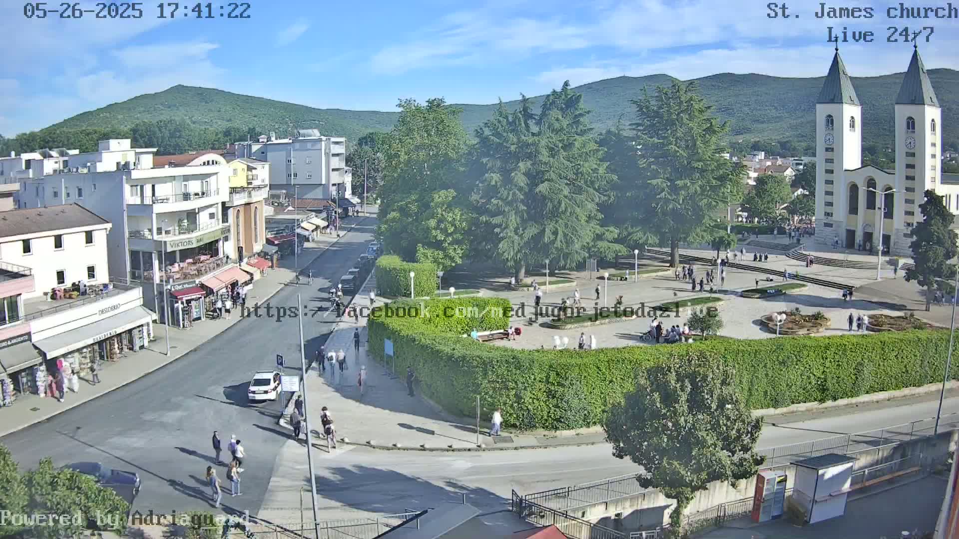 A town square with a large church, surrounded by buildings and green spaces, under a mostly sunny sky.