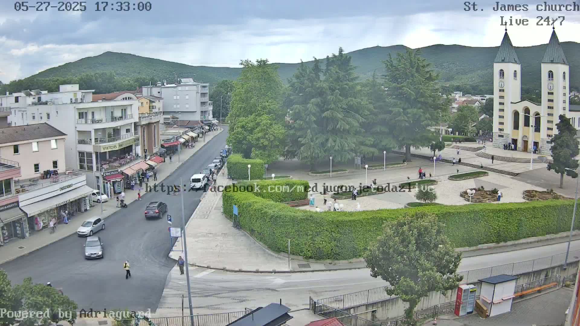 A town square with a church, shops, and cars, under a partly cloudy sky.