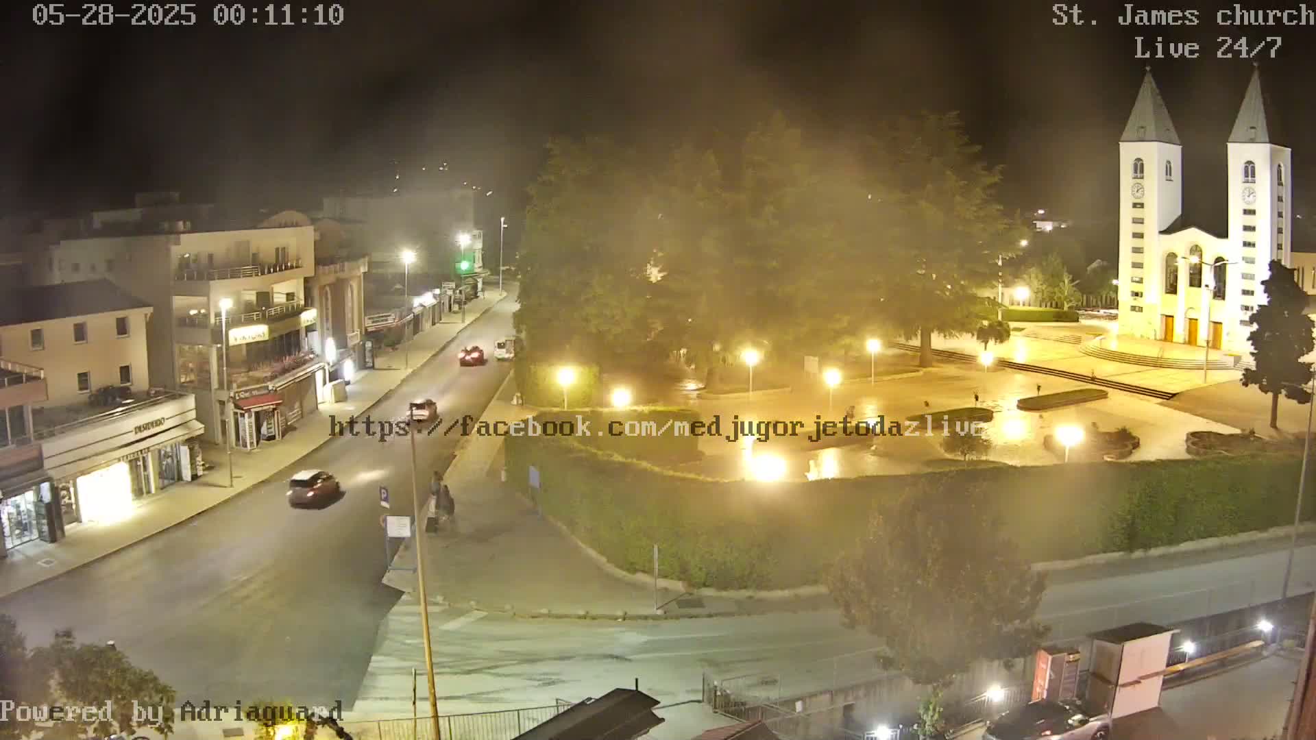 A nighttime, hazy view of a town square with a church, buildings, and cars on the road.