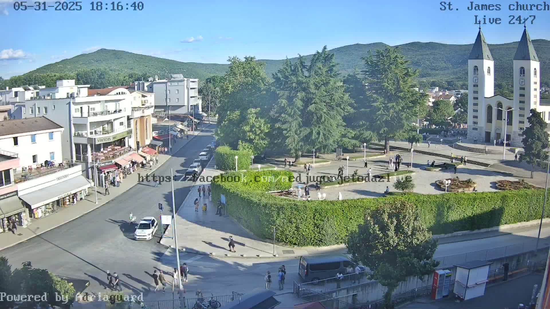 A sunny day overlooking a town square with a church, green spaces, buildings, and pedestrians.