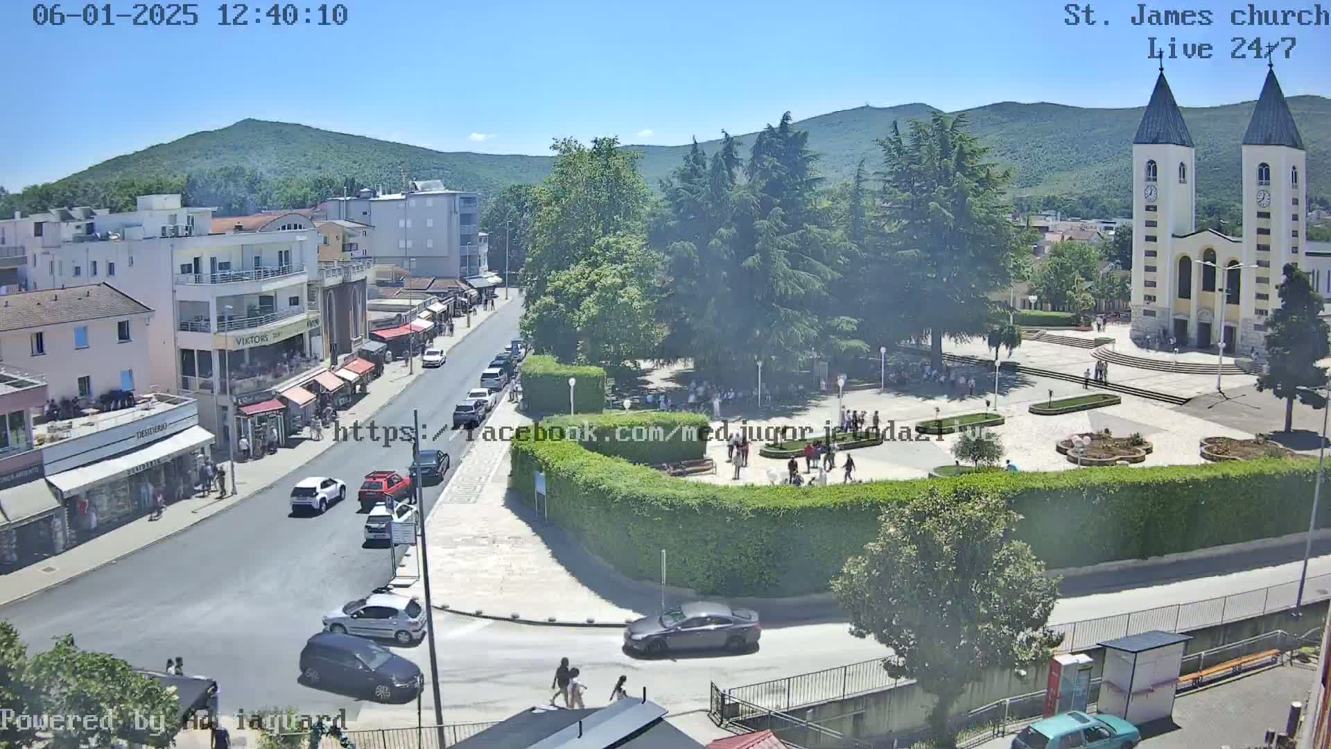 A sunny day reveals a town square with a church, surrounded by buildings, parked cars, and people walking, all under a clear blue sky.