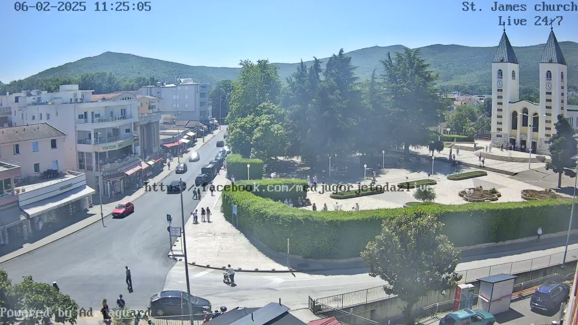 A sunny, high-angle view of a town square with a church, buildings, and people, surrounded by lush greenery and mountains in the background.