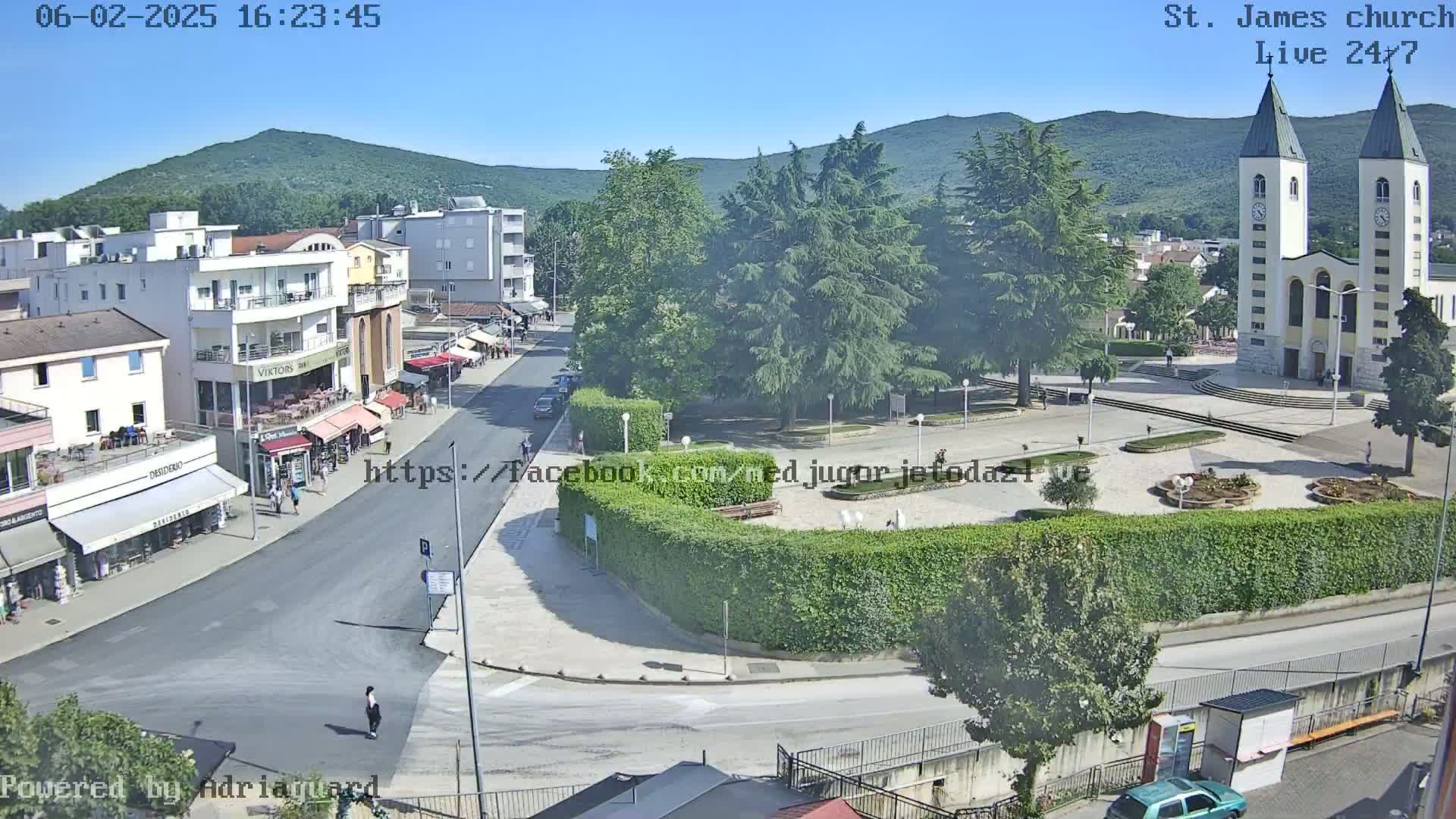 A sunny, clear day overlooking a town square with a church, buildings, and a street, surrounded by greenery and mountains in the distance.