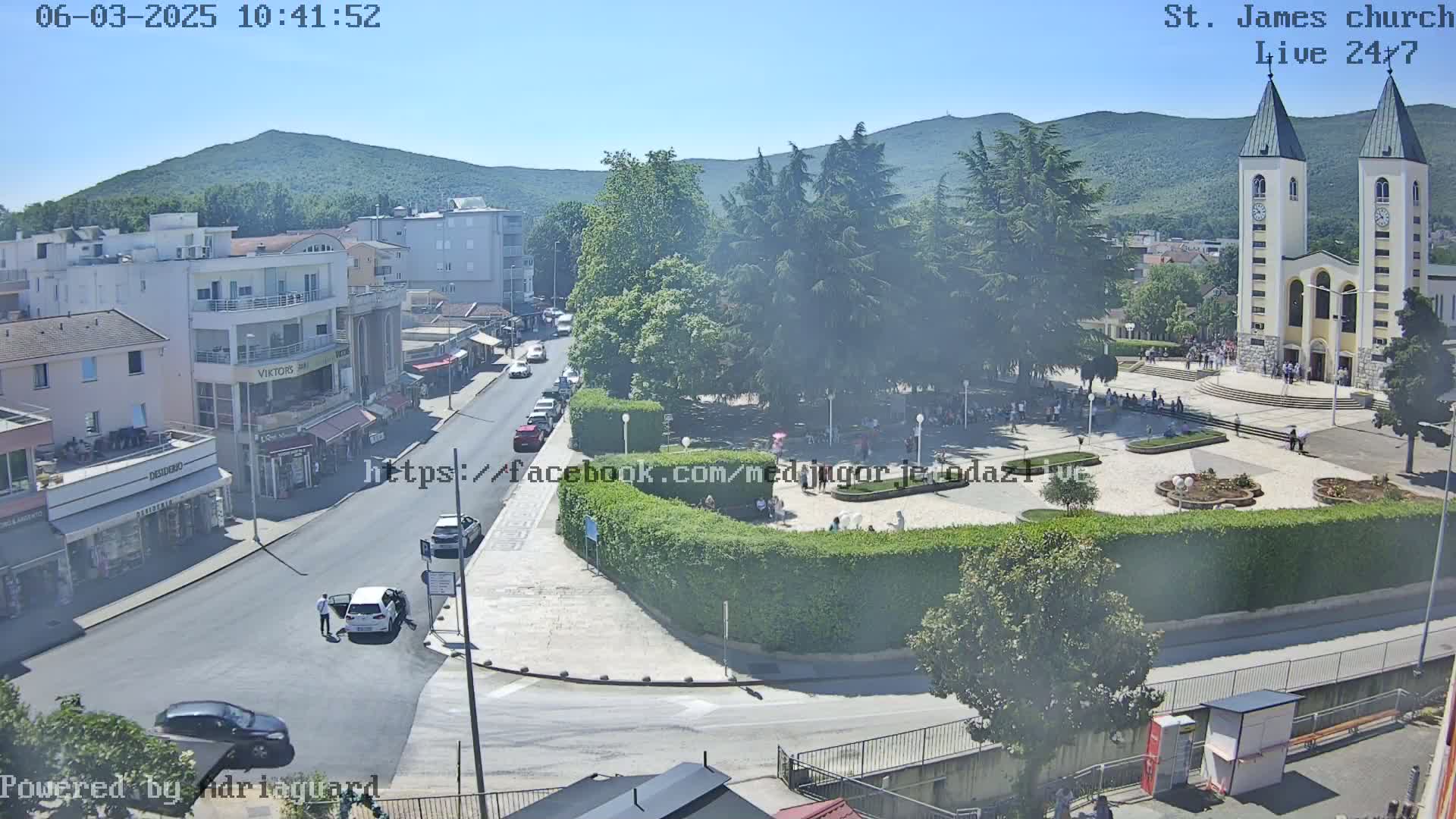 A sunny day view of a town square with a church, buildings, parked cars, and people; a green hill is in the background.