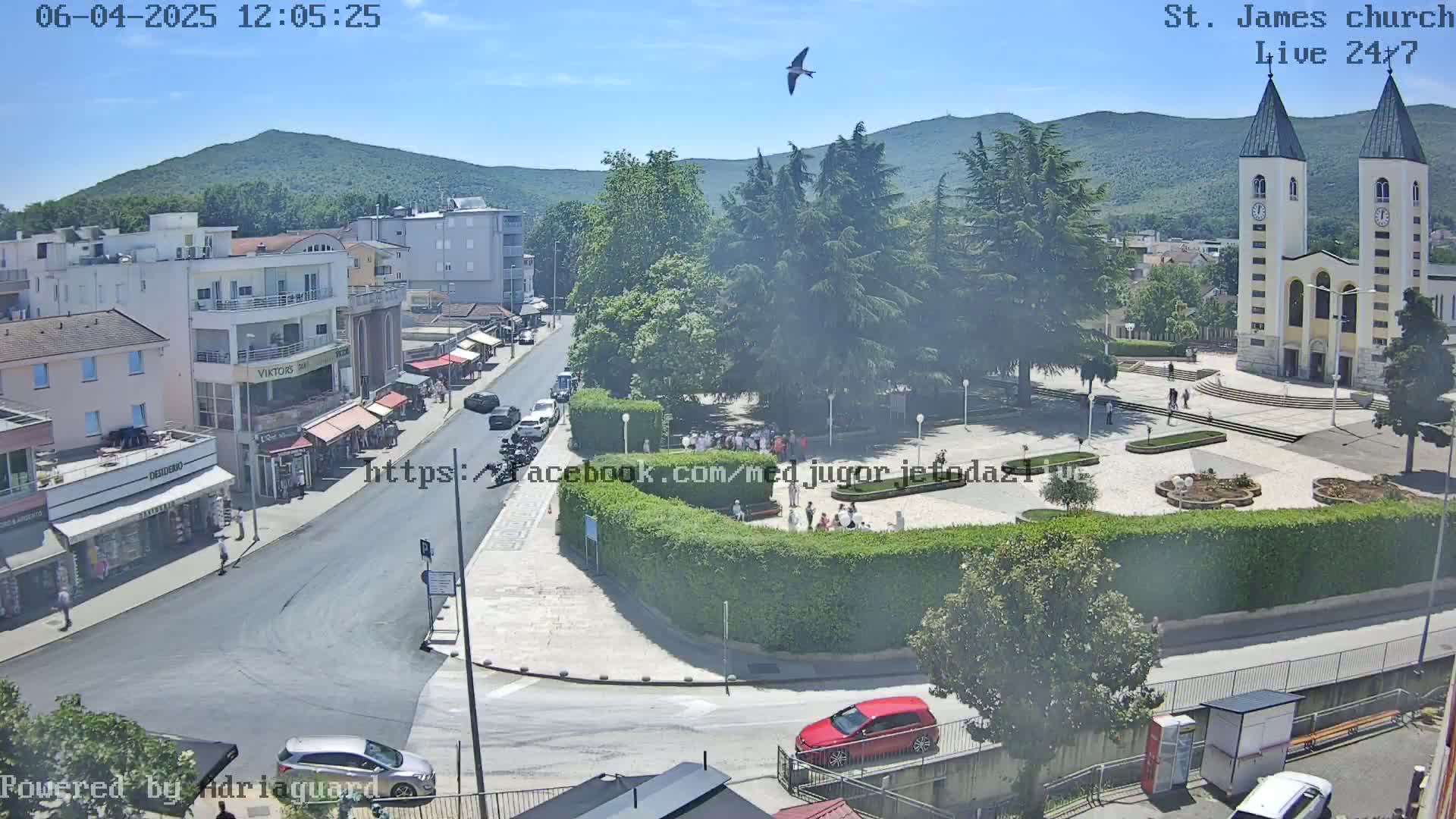 A high-angle, sunny view of a town square with a church, people, cars, and lush green trees, under a clear blue sky.