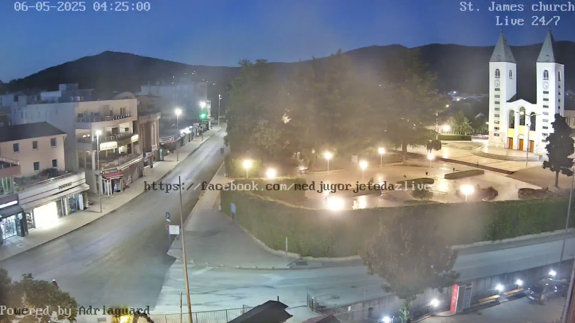 A nighttime, slightly hazy view of a town square featuring a large church, with a road leading to it from the left, and buildings lining the street.