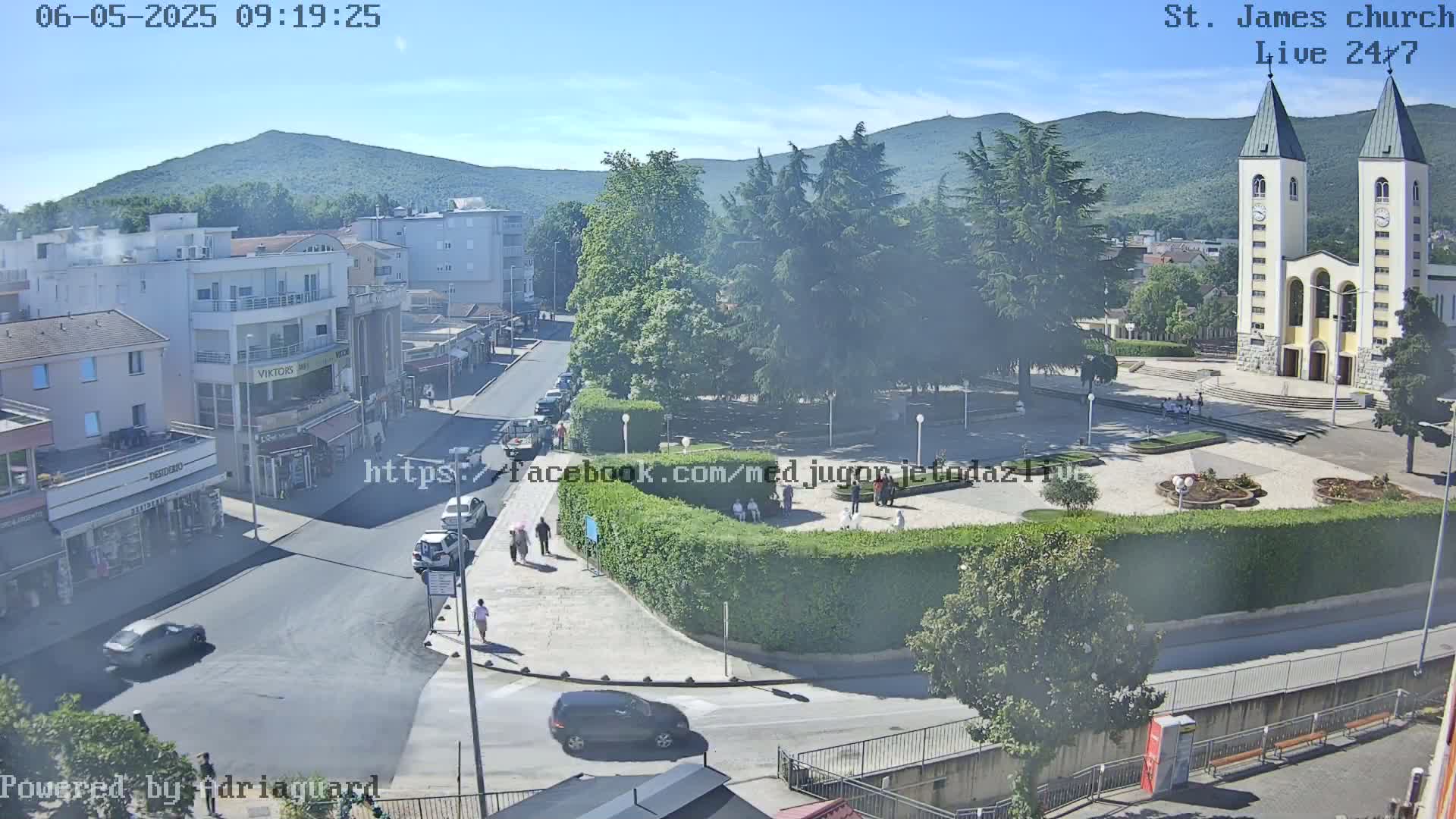 A high-angle, long shot of a town square on a sunny day, with a large church in the background and several cars parked along the street.