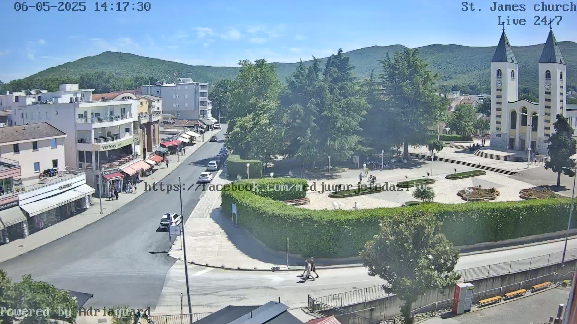 A sunny day reveals a town square with a church, shops, and people, situated next to a road and backed by green hills.