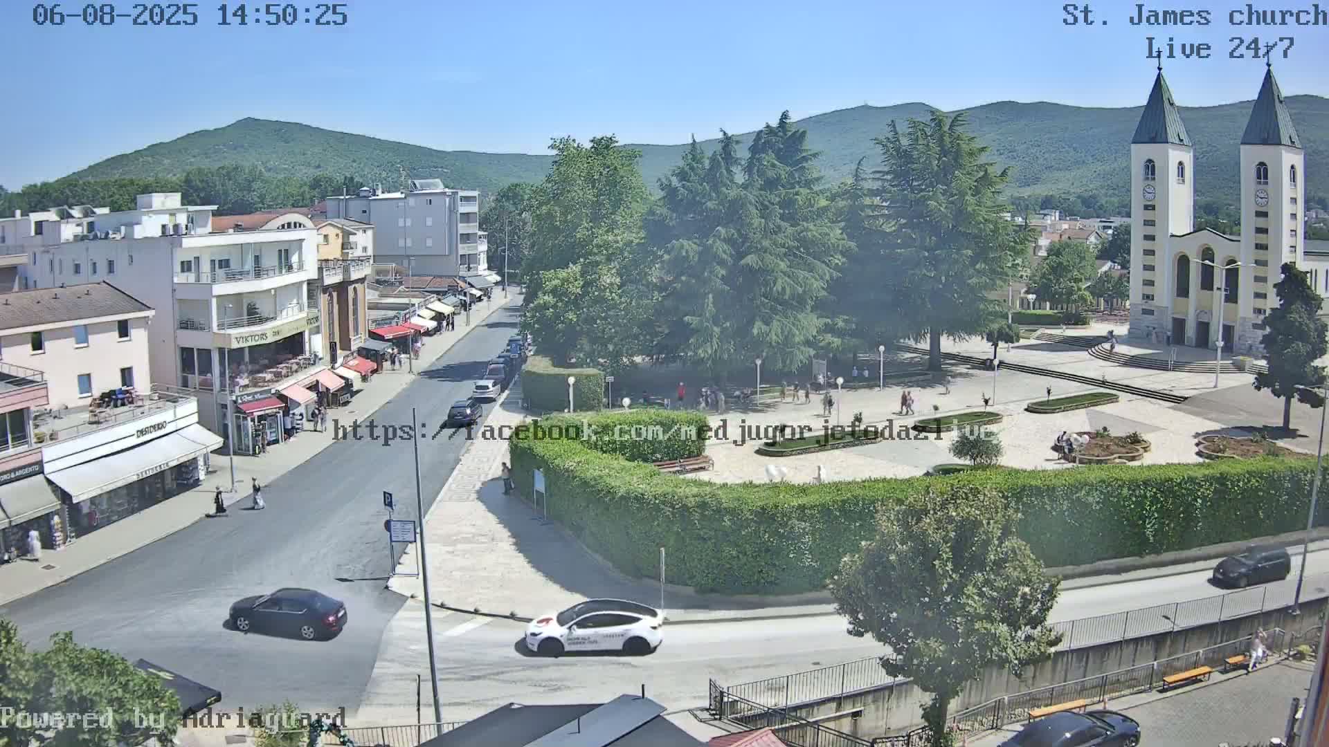 A sunny day in a town square with a large church, featuring several buildings, cars, and pedestrians; lush green trees surround a meticulously landscaped area.